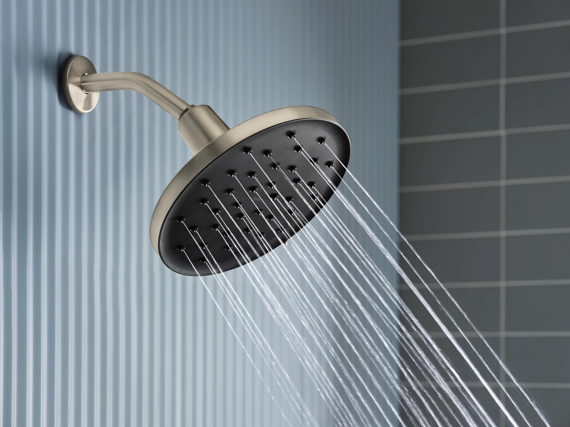 Brushed nickel rainshower head with curved arm mounted on tile wall, water streams visible from perforated face.