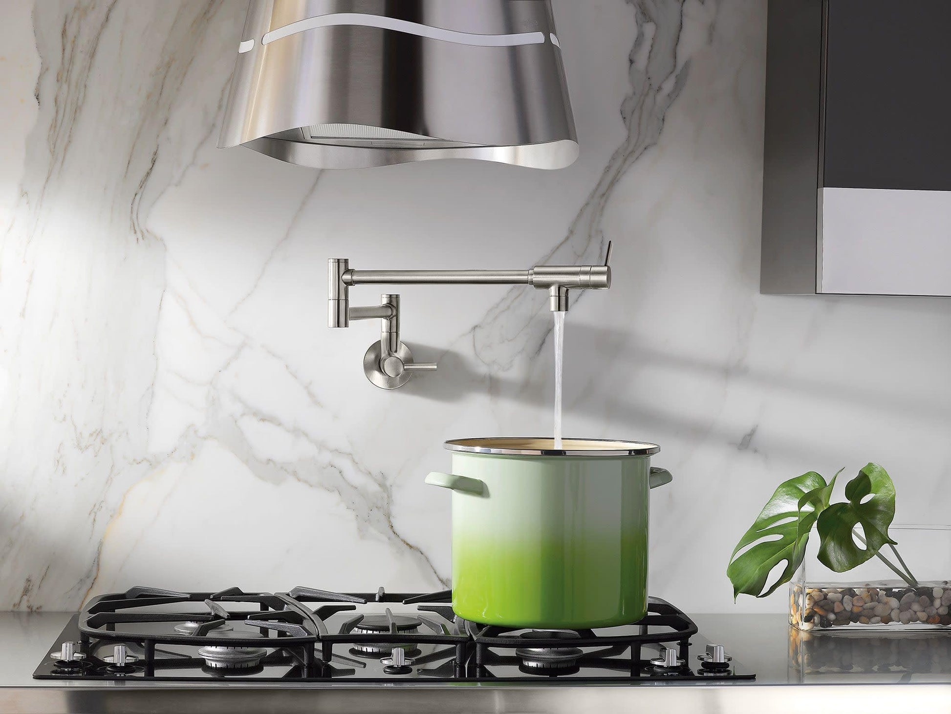 Stainless steel wall-mounted pot filler faucet with double-jointed swing arm positioned above stovetop with green pot.