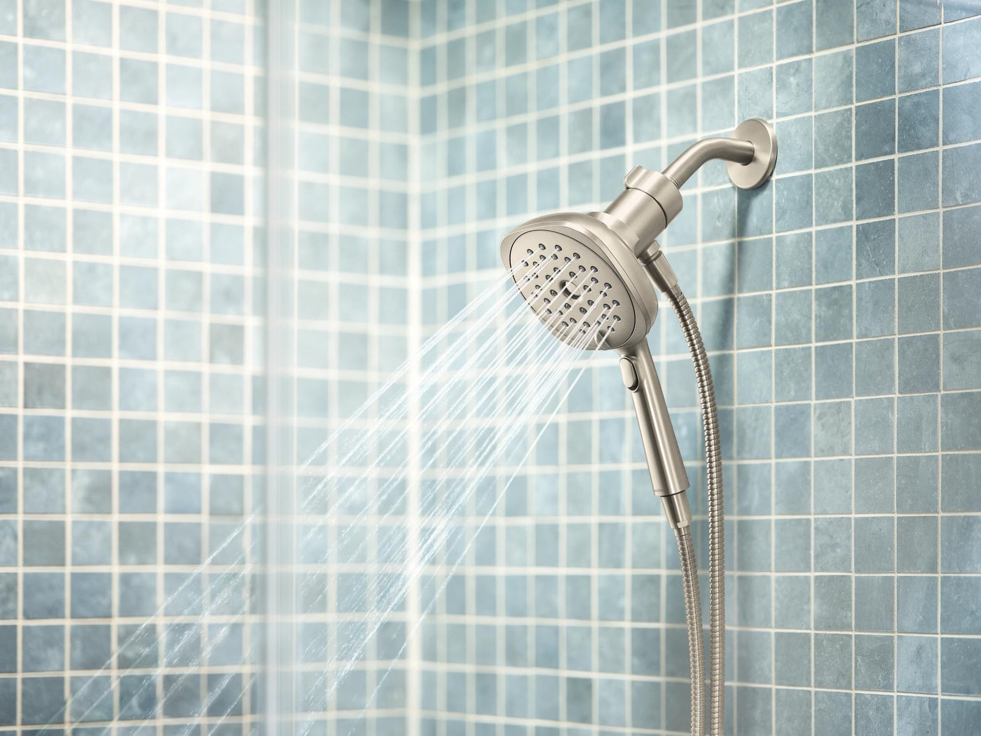 Brushed nickel handheld showerhead with square spray pattern mounted on tile wall with connected hose.