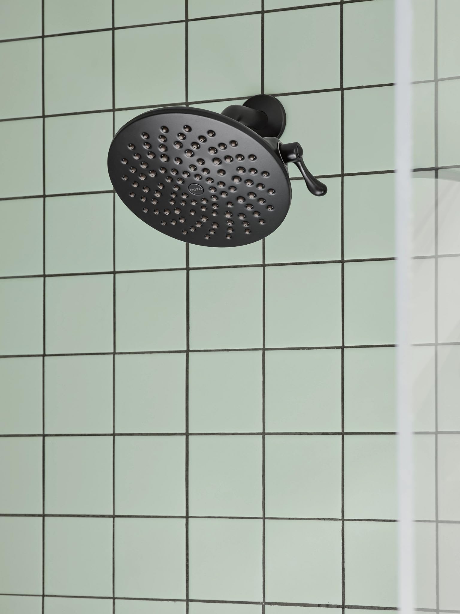 Matte black round showerhead with multiple spray holes mounted on tiled shower wall with control lever.