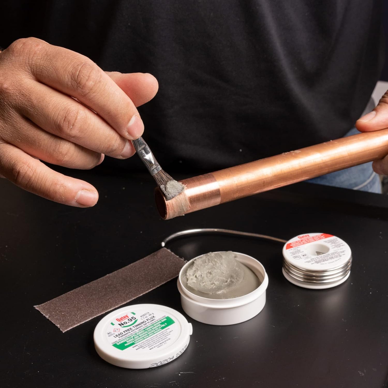 Person applying tinning flux with brush to copper pipe, with containers of flux and solder visible below.