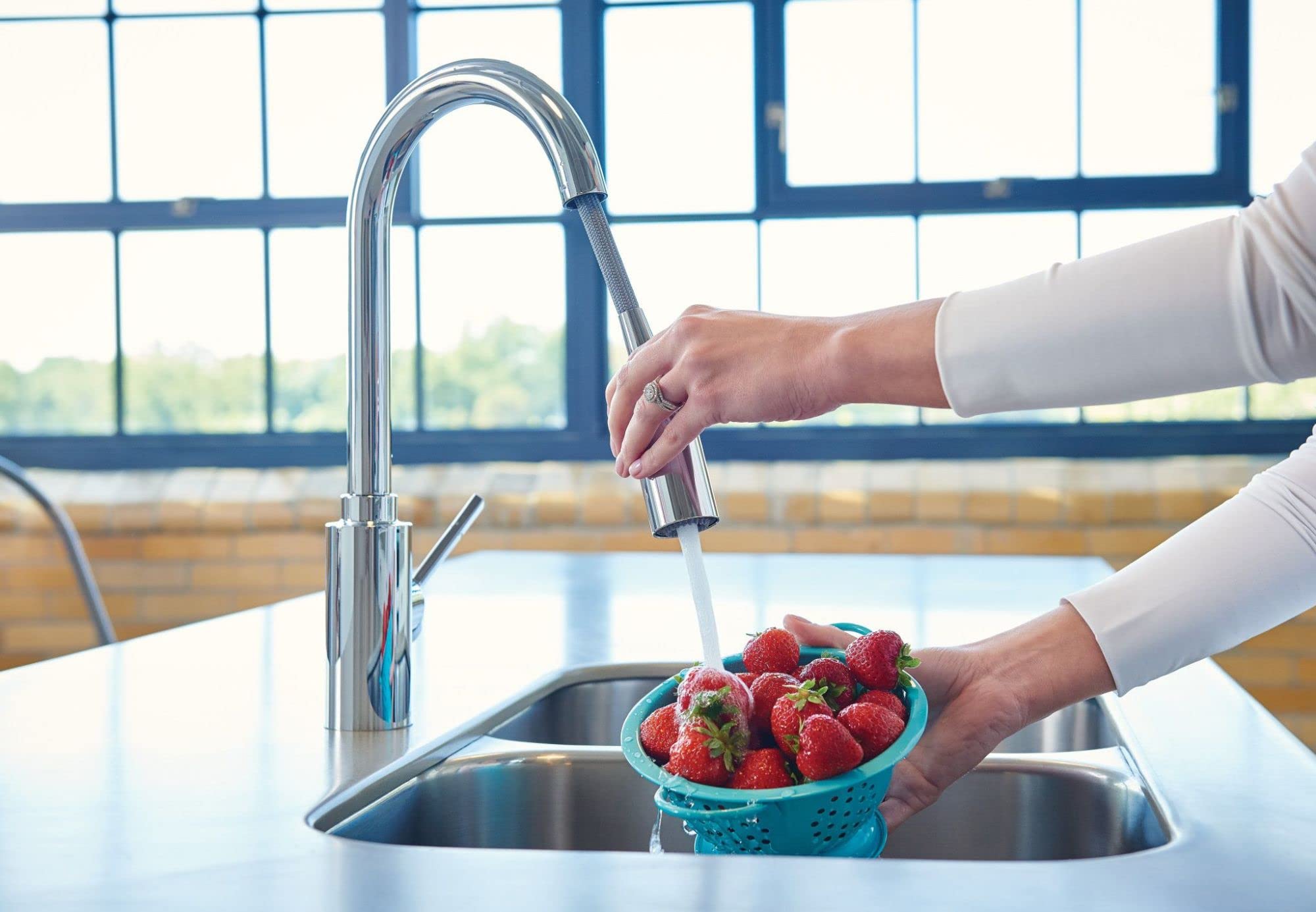 Chrome kitchen faucet with pull-down sprayer over double sink basin, hand rinsing strawberries.
