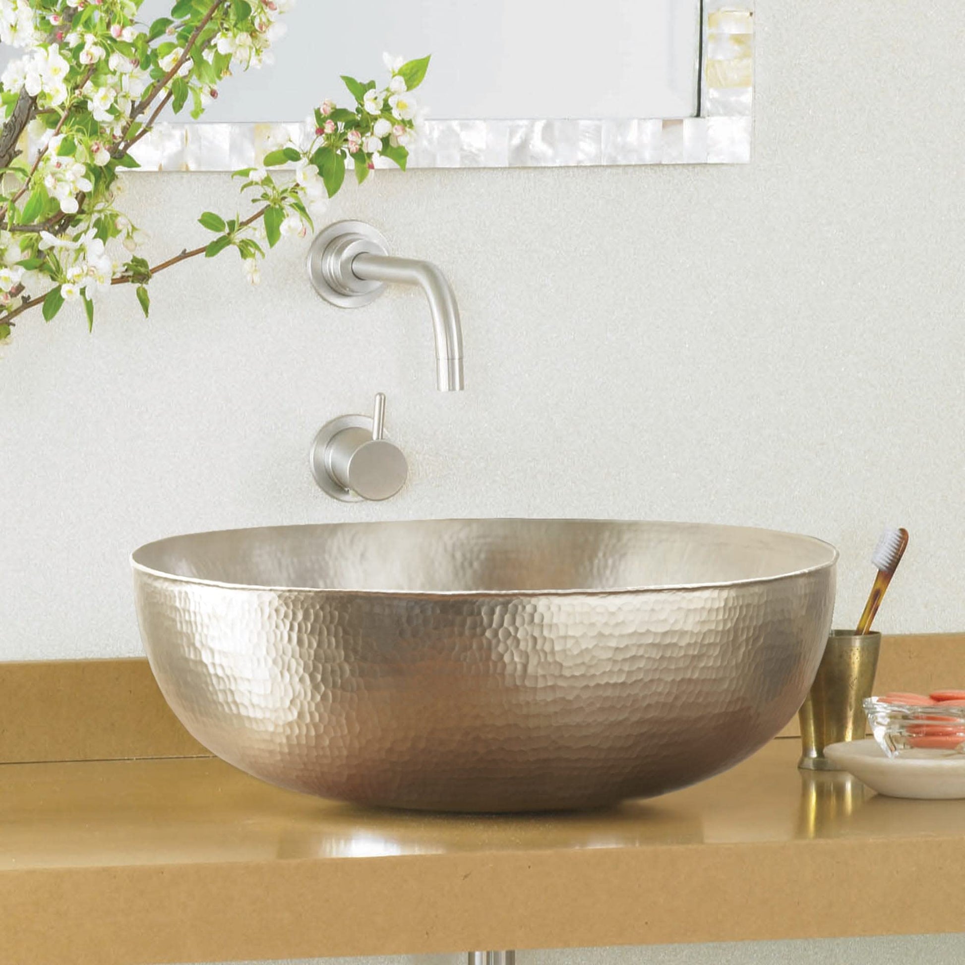 Oval metallic bathroom sink with hammered finish surface and wall-mounted faucet in brushed nickel.