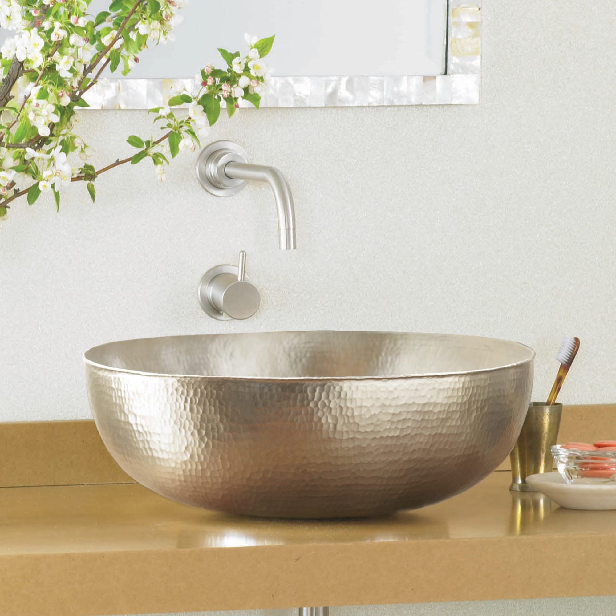 Oval metallic bathroom sink with hammered finish surface and wall-mounted faucet in brushed nickel.