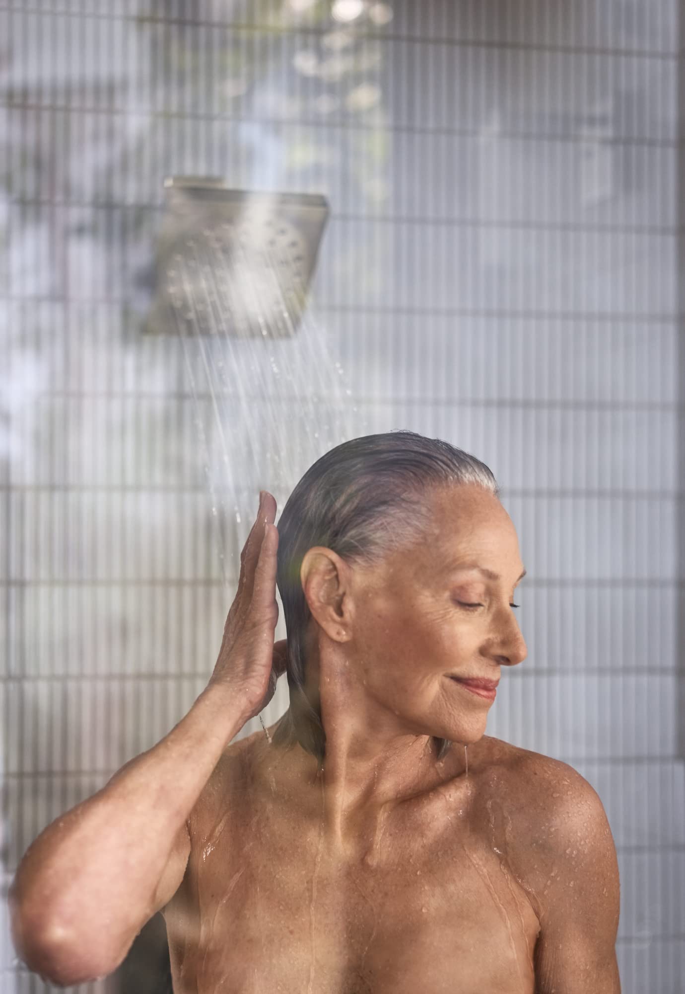Brushed nickel shower head with angled design spraying water over a person's head in a tiled shower.