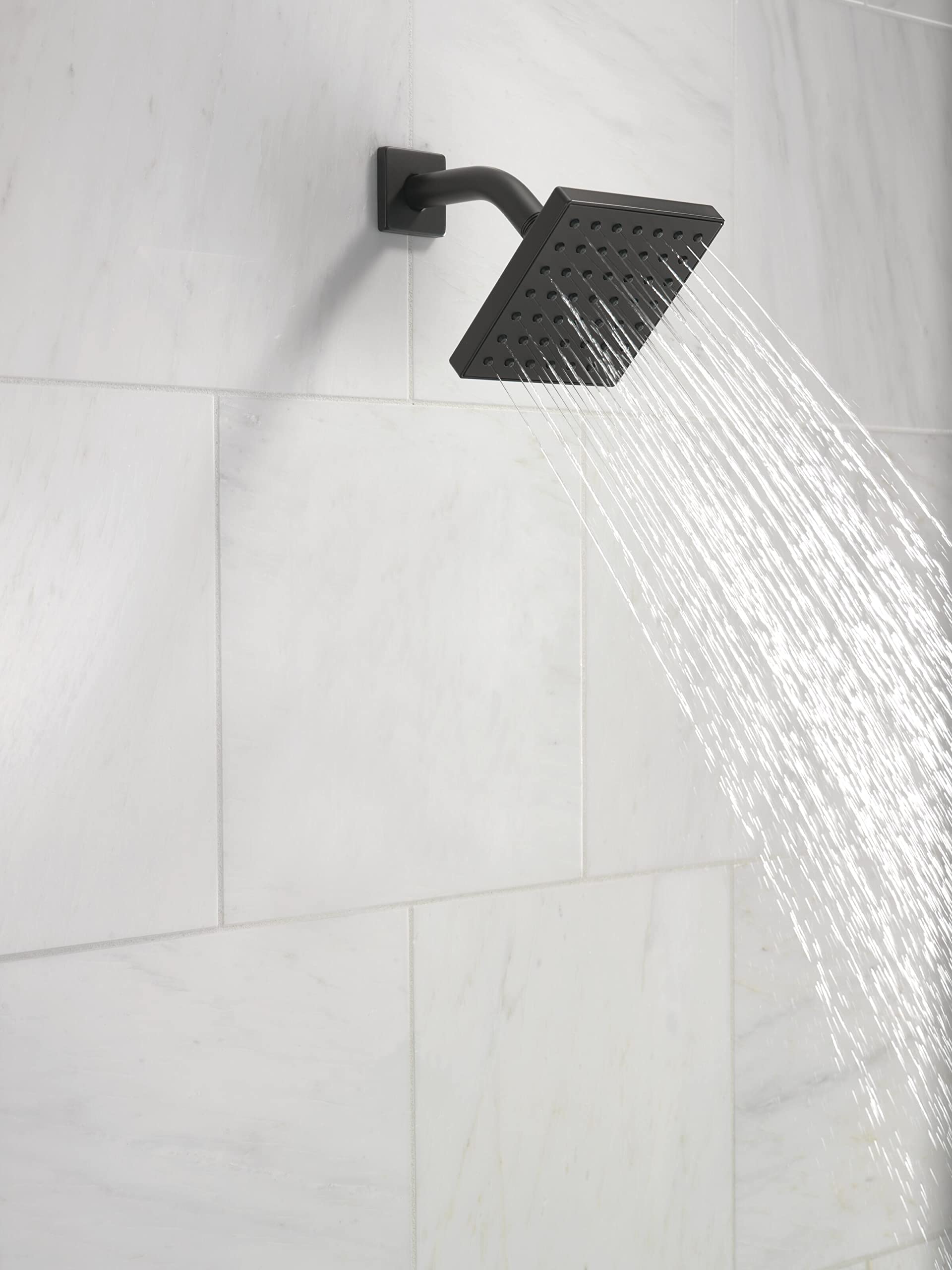 Square matte black shower head mounted on white tile wall with water flowing from perforated head.