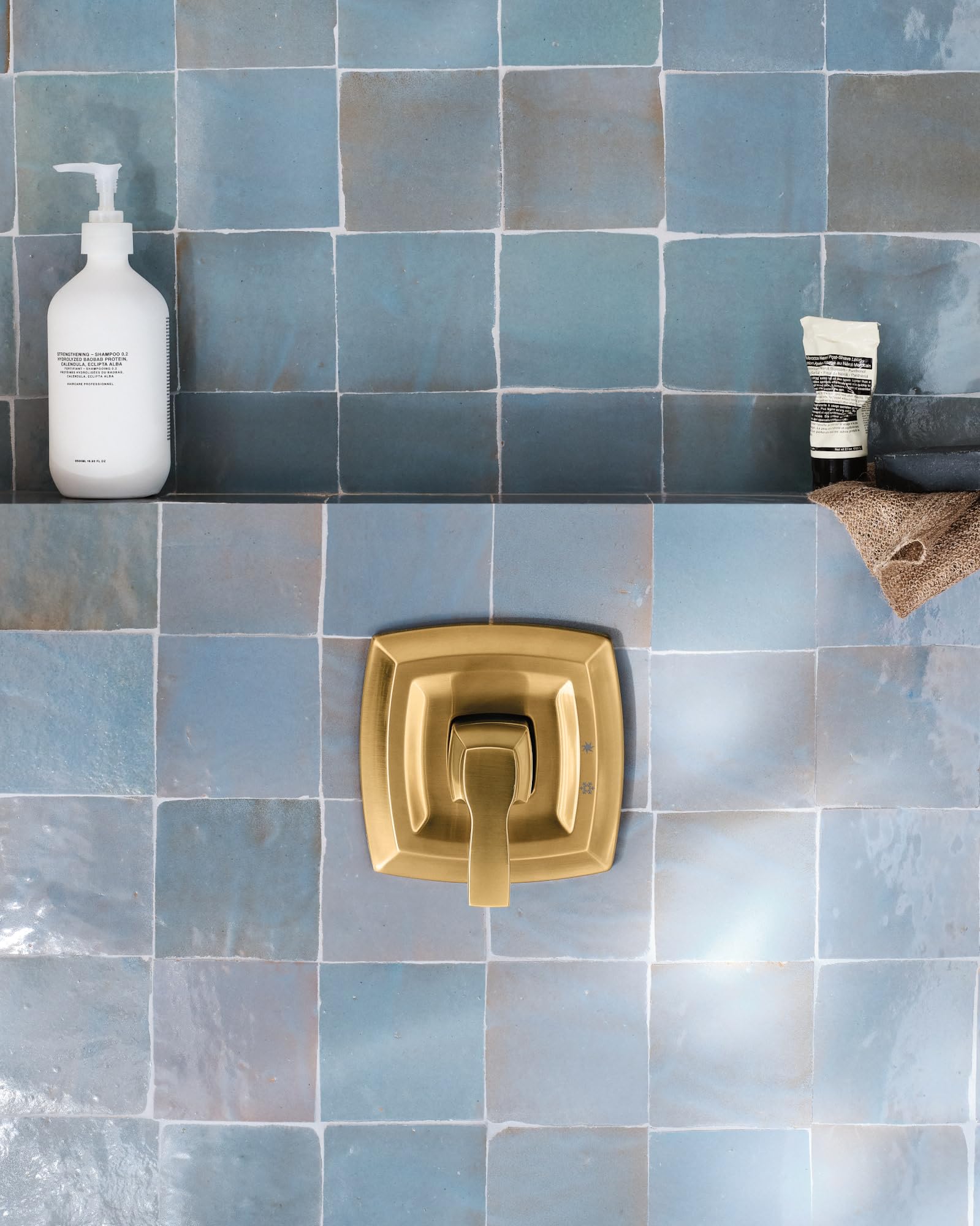 Gold square shower valve trim with single lever handle mounted on blue tile wall.