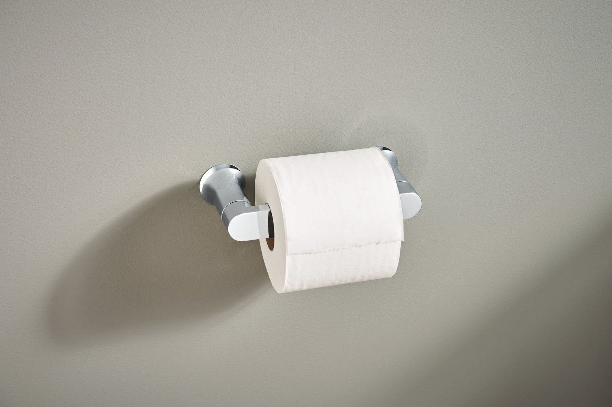 Chrome toilet paper holder with pivoting arms mounted on beige wall holding white roll.
