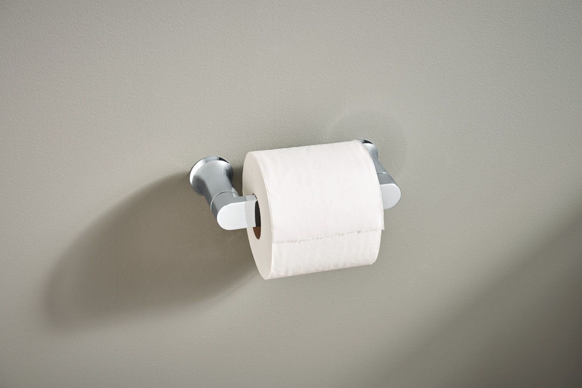 Chrome toilet paper holder with pivoting arms mounted on beige wall holding white roll.