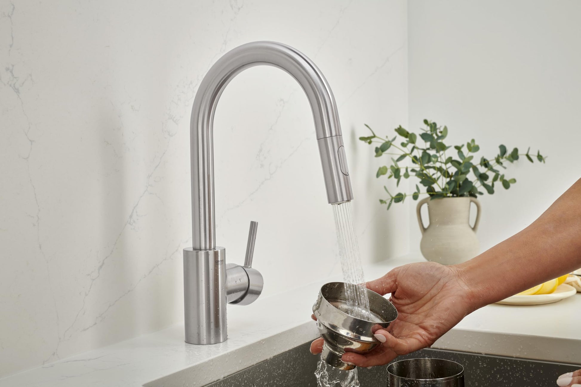 Stainless steel pull-down bar faucet with curved spout dispensing water into a cup held in a hand over a dark countertop.