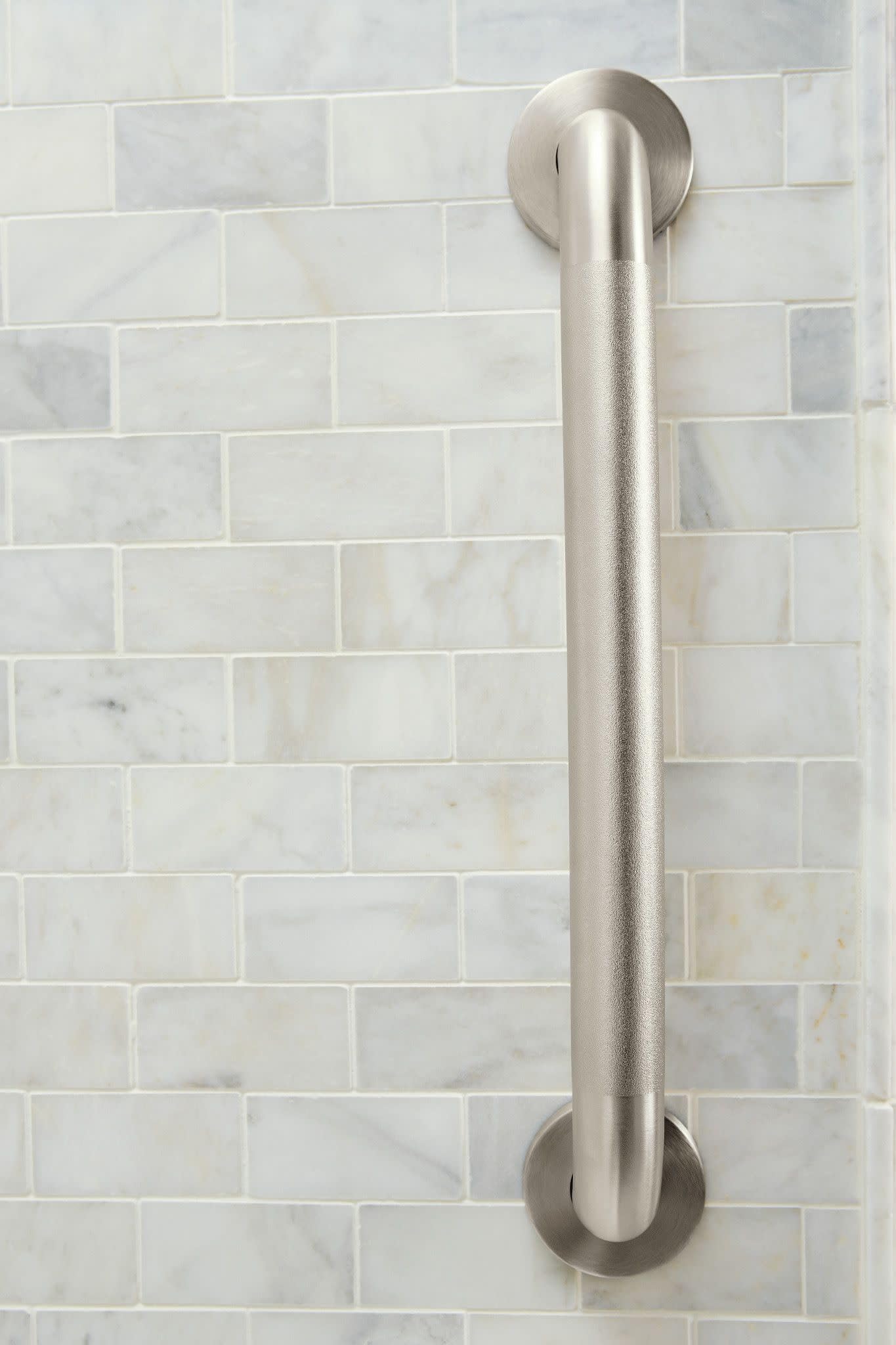 Brushed nickel grab bar with peened finish mounted vertically on marble subway tile wall.