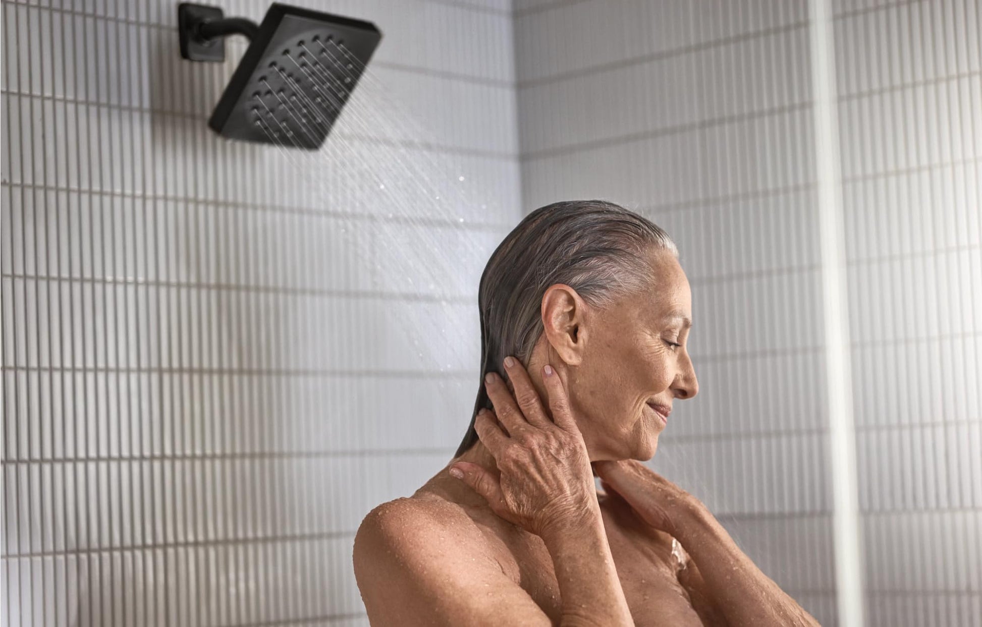 Matte black square showerhead mounted on white tile wall with water flowing, person showering below.