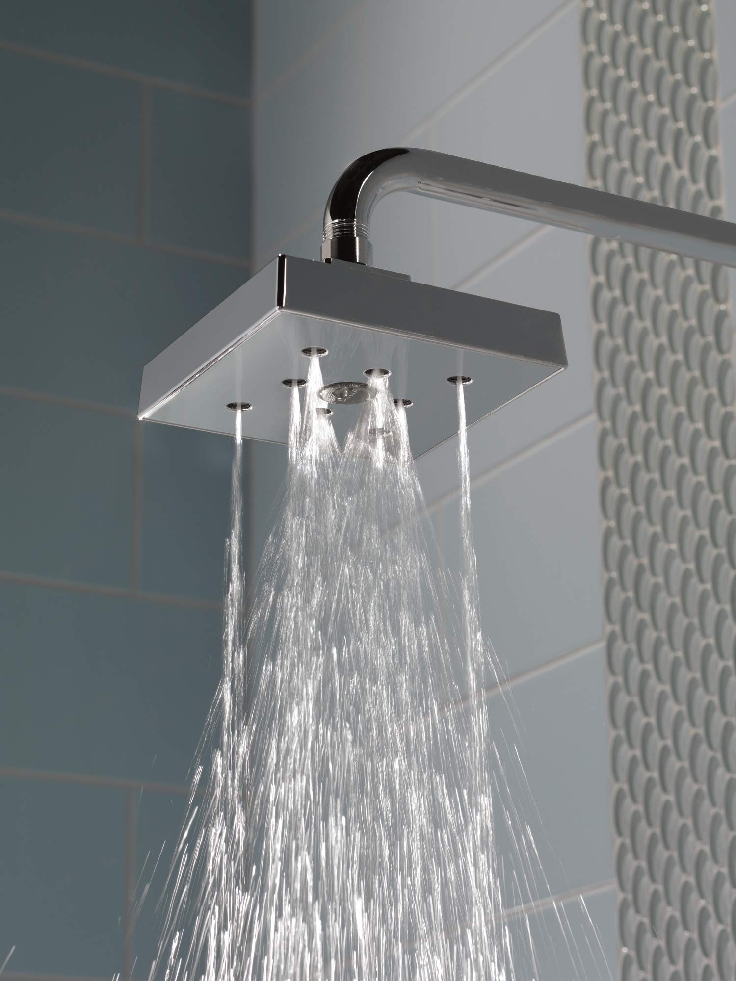Black stainless steel shower arm with square head and curved connection, water flowing downward against tiled bathroom wall.