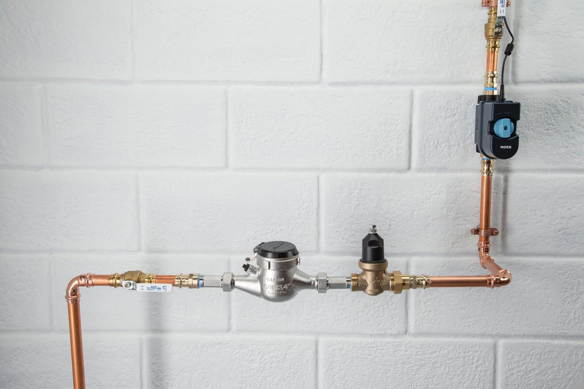 Black Moen Flo smart water monitor device mounted on copper water pipe with shutoff valve against white brick wall.