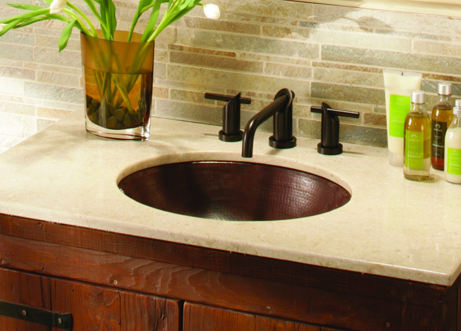 Oval hammered copper bathroom sink with antique patina, mounted in beige countertop with black faucet.