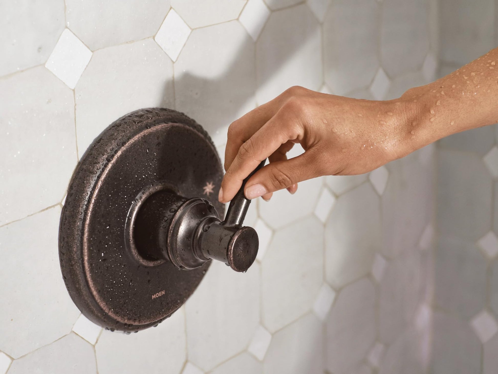 Oil rubbed bronze single-handle tub and shower trim with circular escutcheon plate mounted on white hexagonal tile wall.