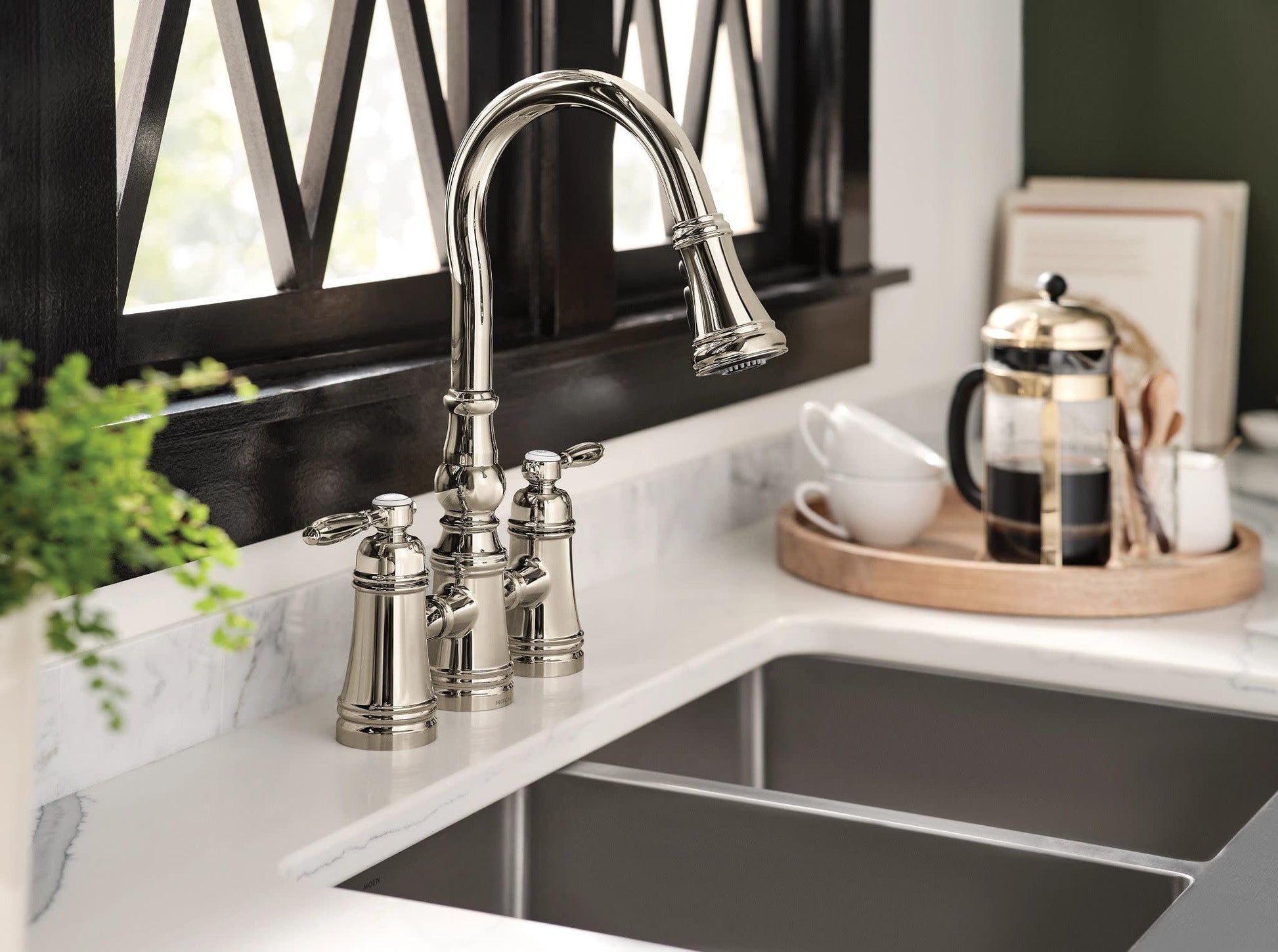 Polished nickel bridge kitchen faucet with curved pulldown spout and two handles installed on white marble countertop.