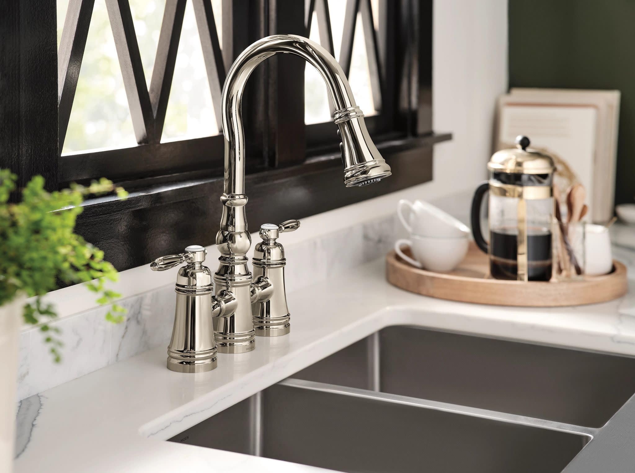 Polished nickel bridge kitchen faucet with curved pulldown spout and two handles installed on white marble countertop.