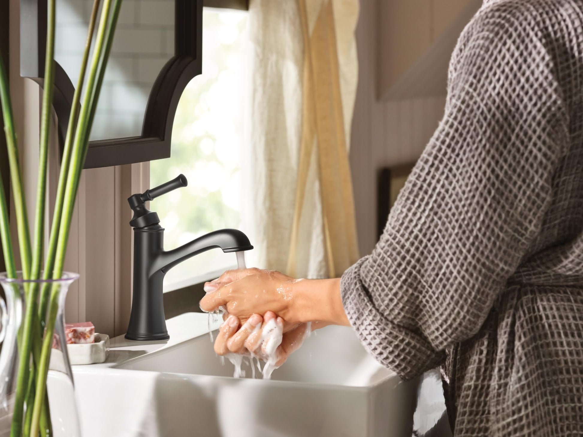 Matte black single-handle bathroom faucet with curved spout installed on white sink with water running over hands.