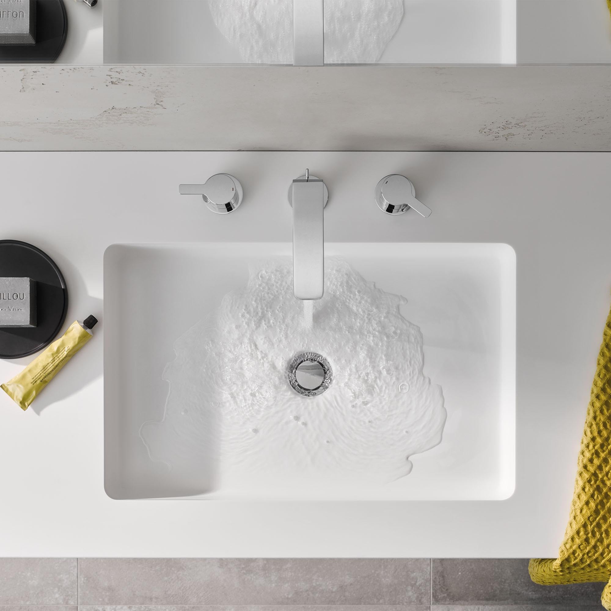 Matte black widespread bathroom faucet with two lever handles mounted above white rectangular sink with water running.