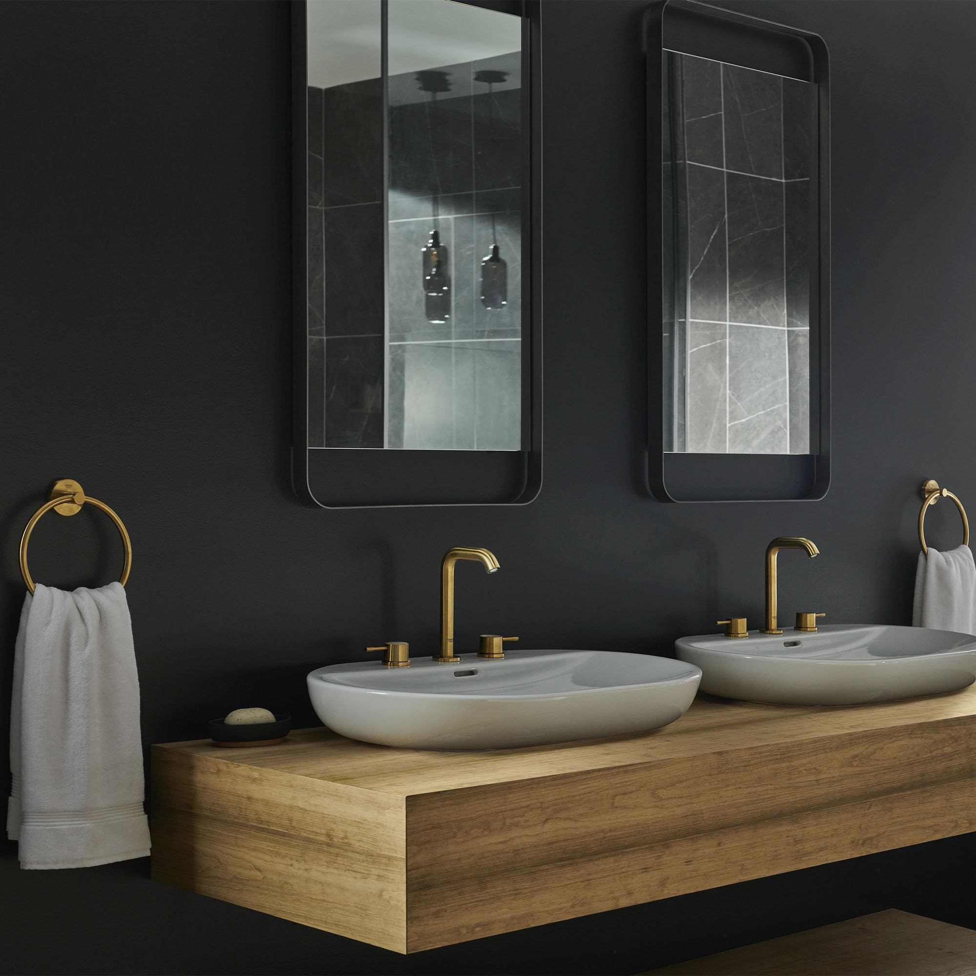 Brushed gold widespread bathroom faucet with two handles mounted on wooden vanity between white oval sinks.