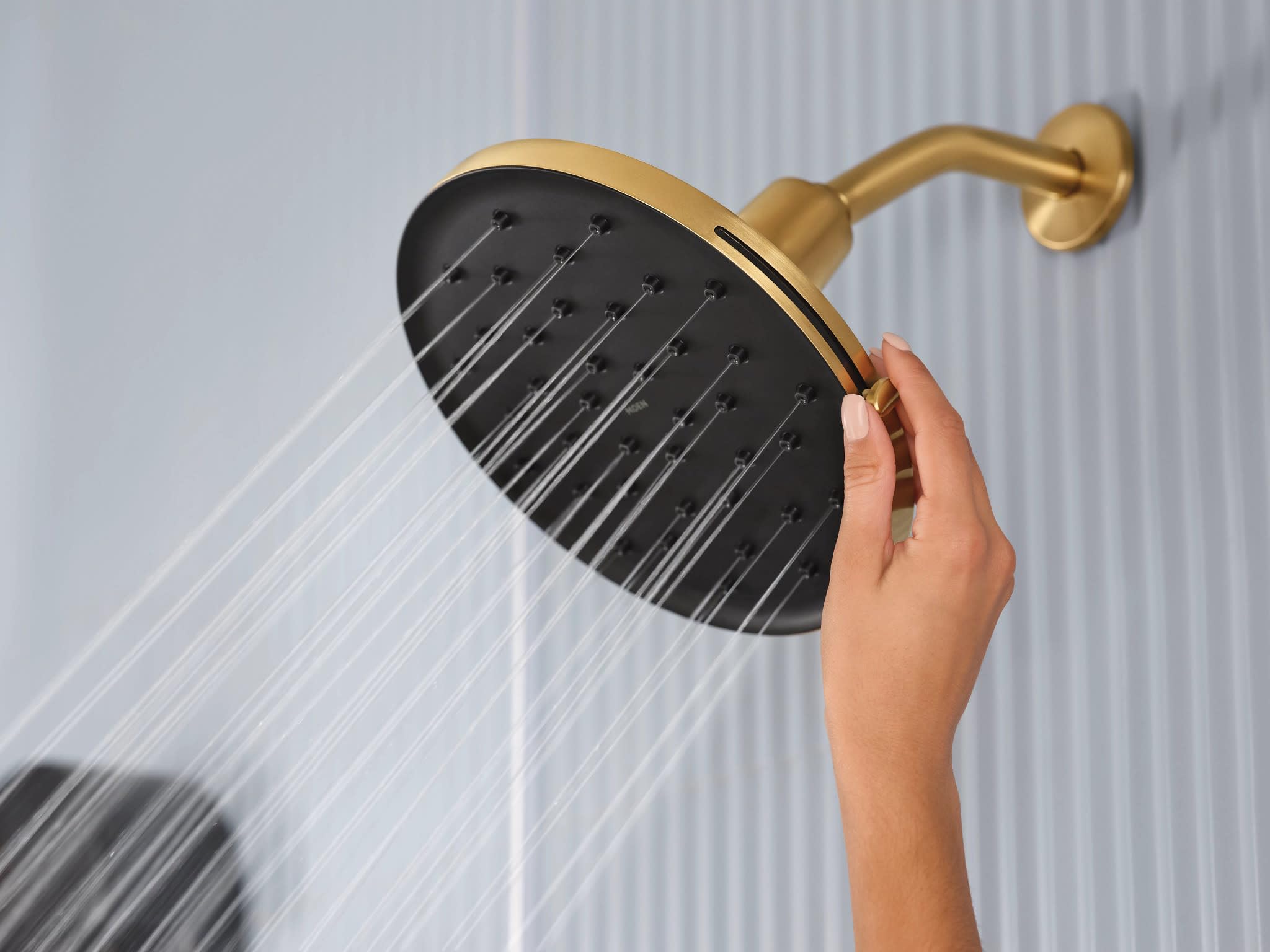 Brushed gold rainfall showerhead with black face and water streams, mounted on curved arm with hand adjusting spray pattern.