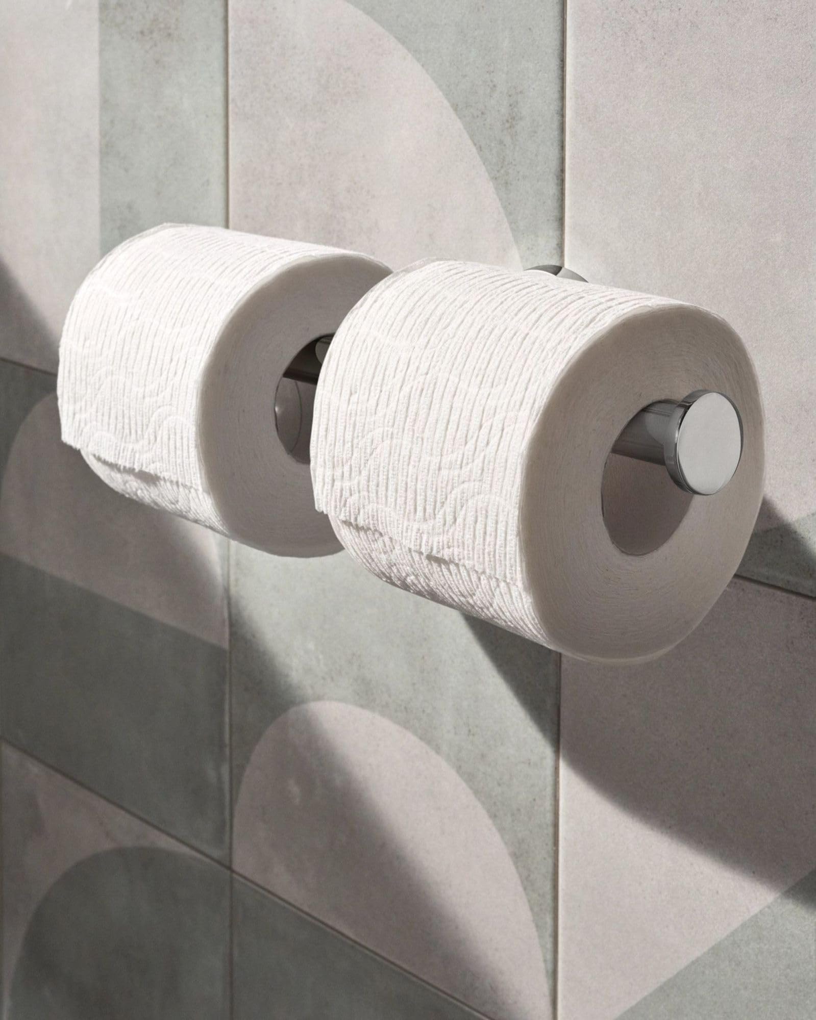 Chrome double toilet paper holder with two white rolls mounted on gray tile wall.