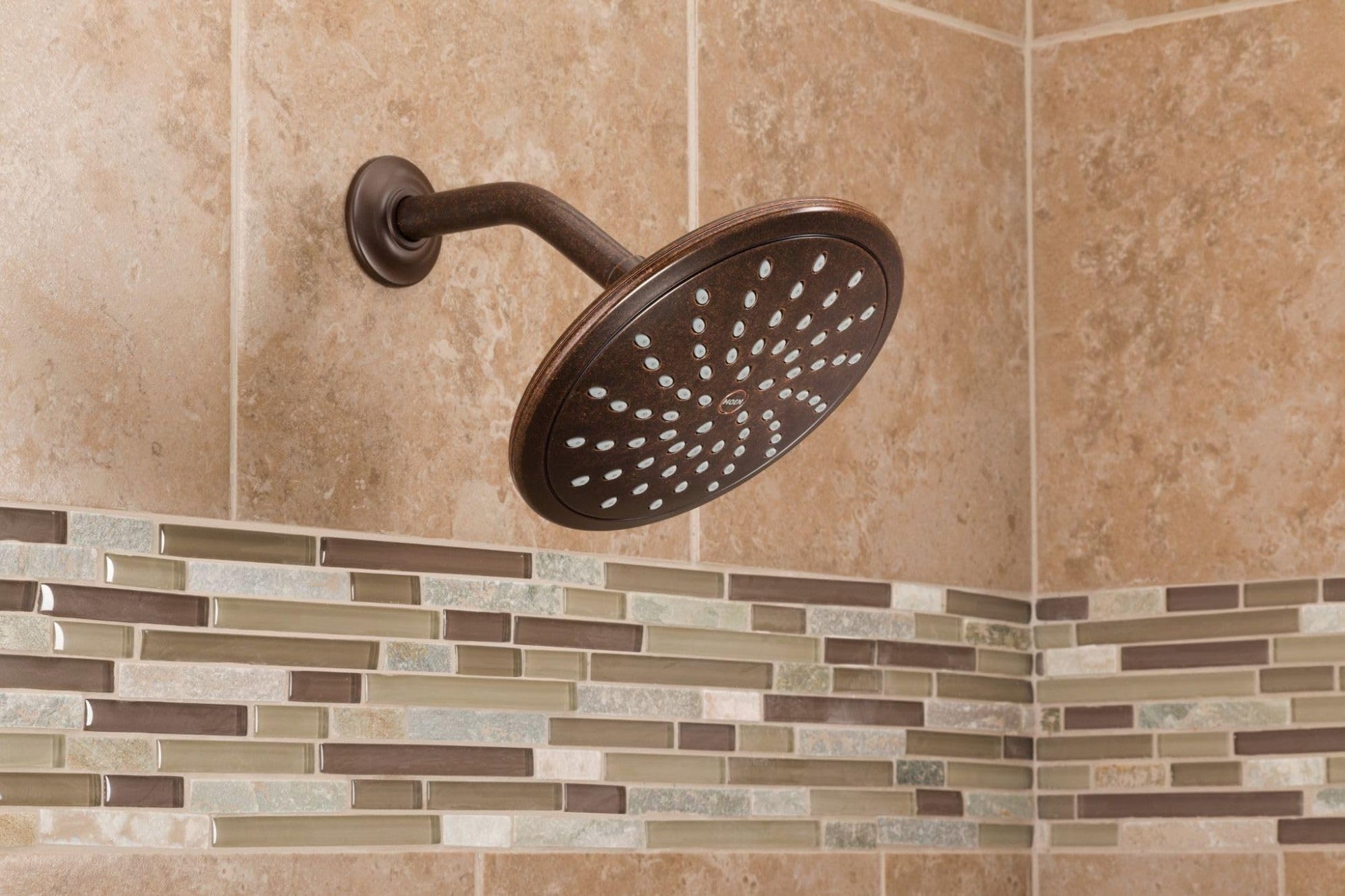 Oil-rubbed bronze round showerhead with spray holes mounted on angled arm against tiled shower wall.