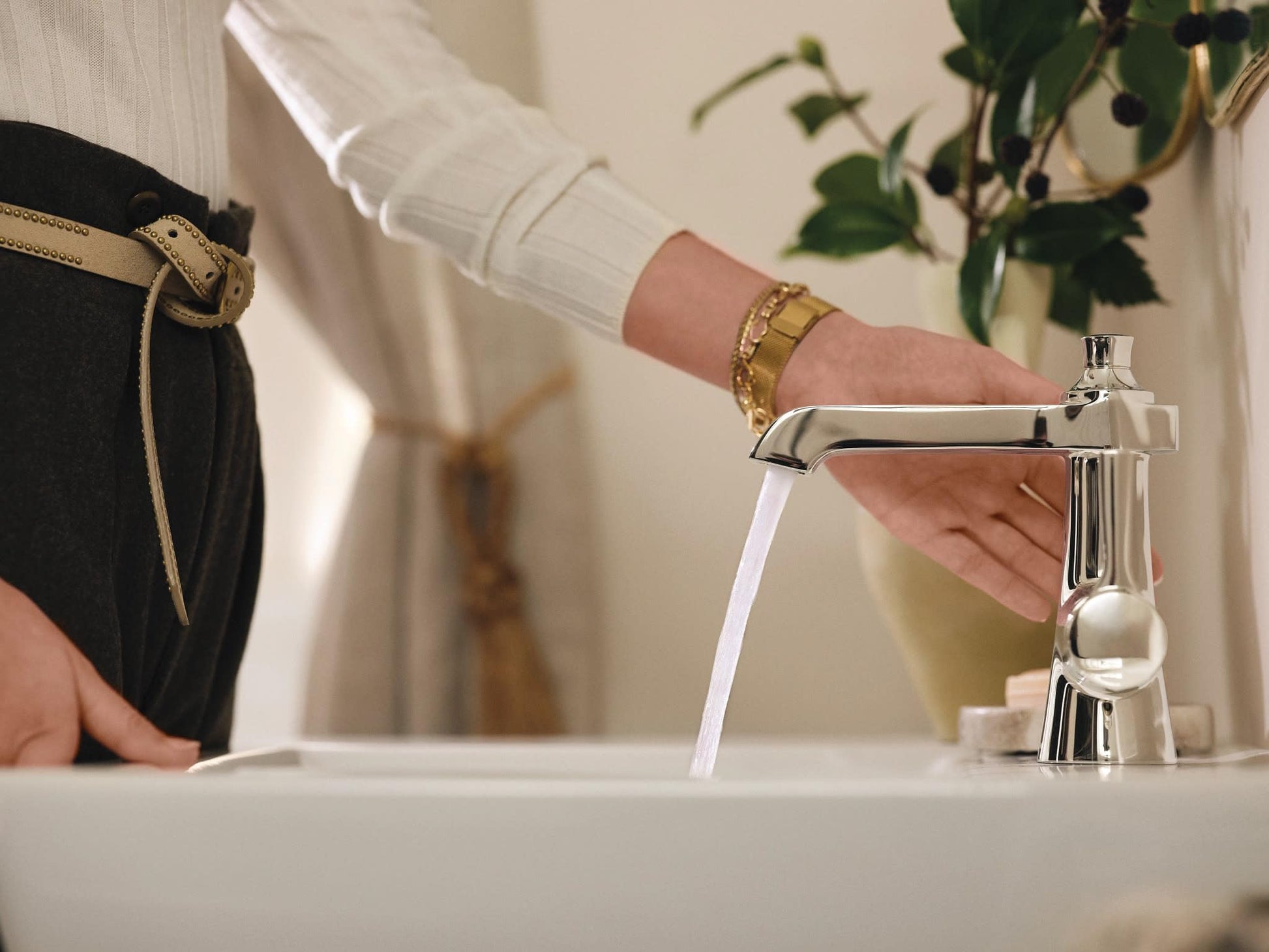 Polished nickel single-handle bathroom faucet with water flowing from spout, person's hand operating faucet over white sink.