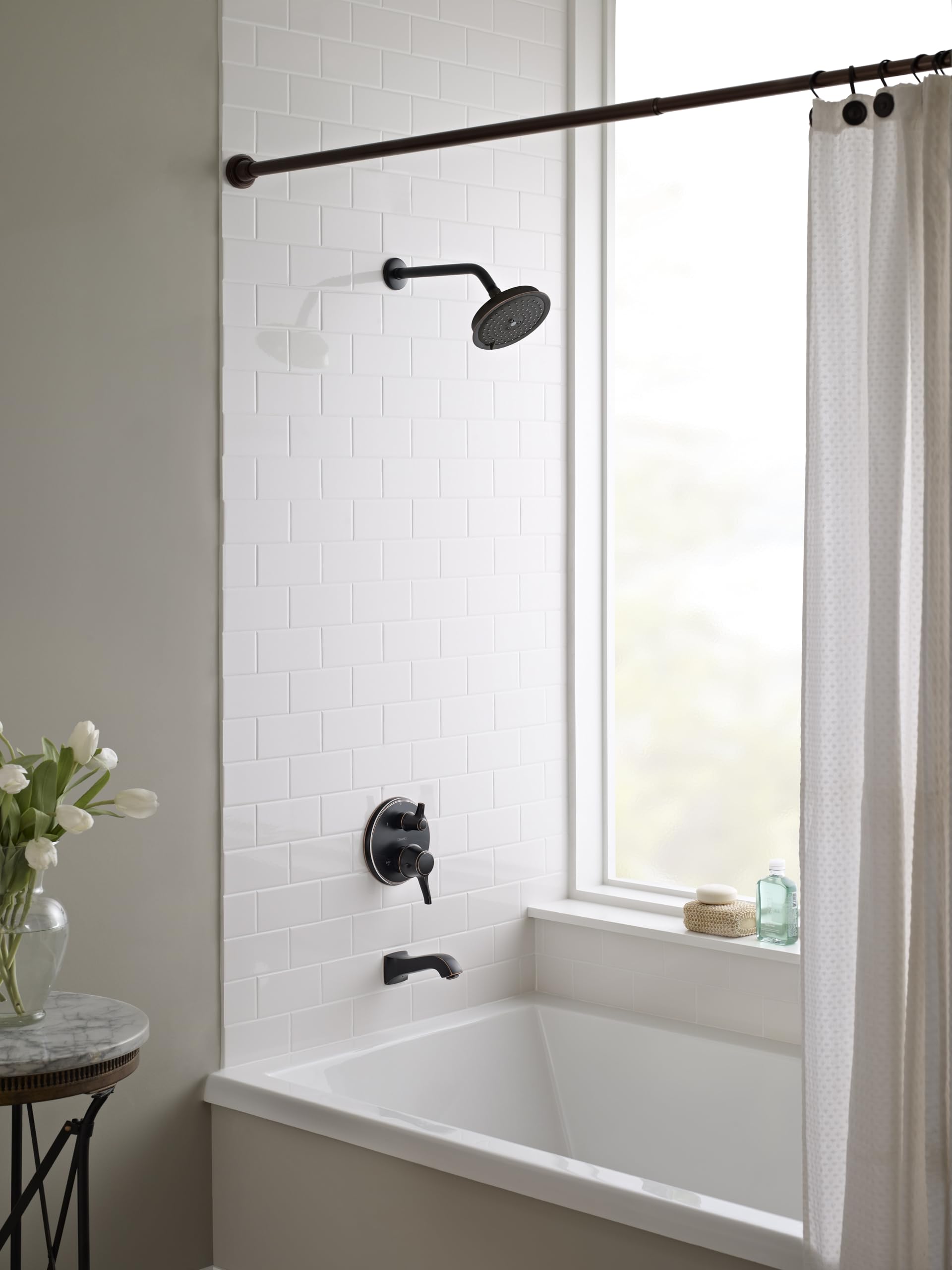 Rubbed bronze shower head with curved arm mounted above white subway tile bathtub surround.