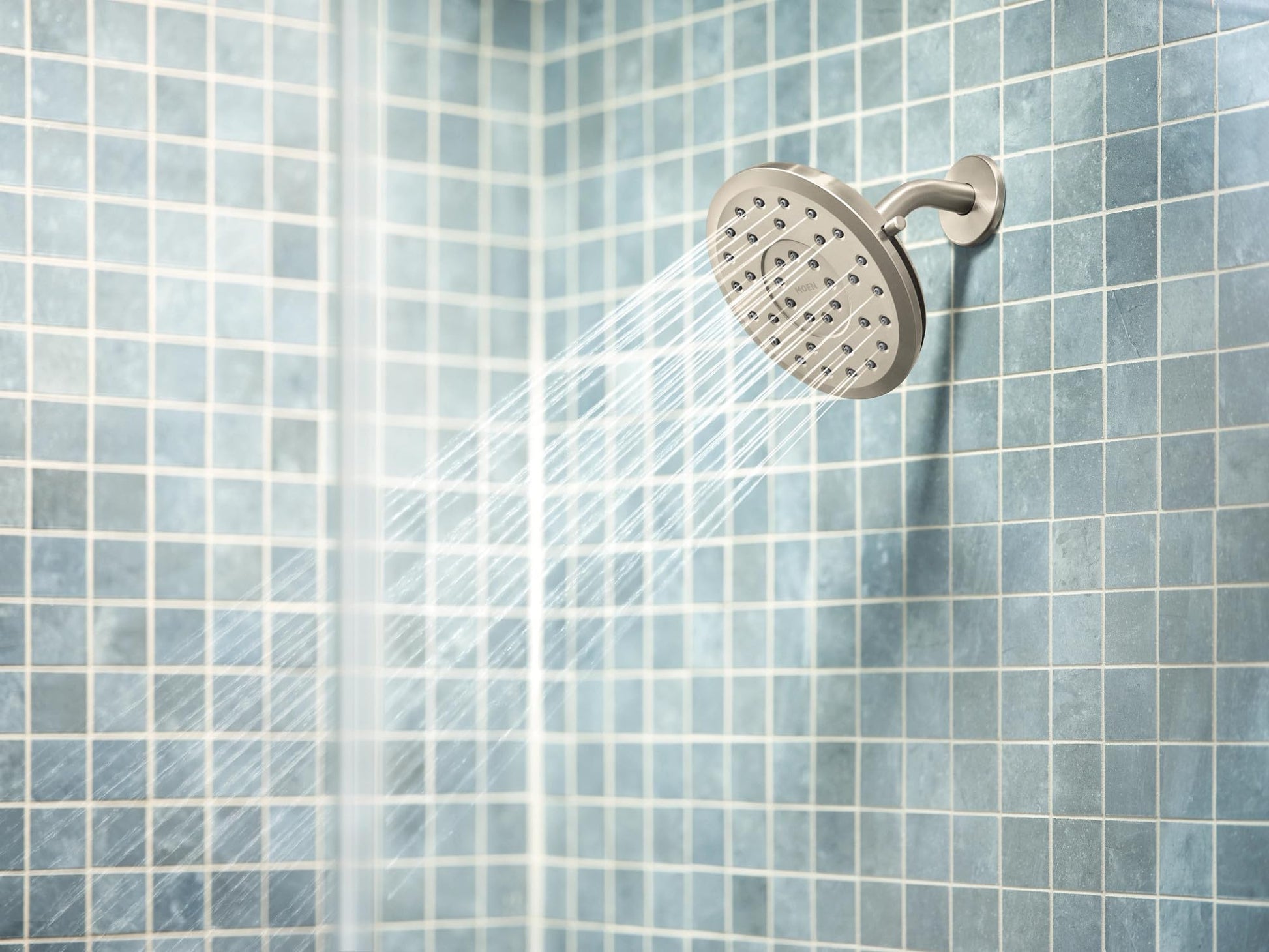 Brushed nickel round rain shower head mounted on tile wall with water streaming downward.