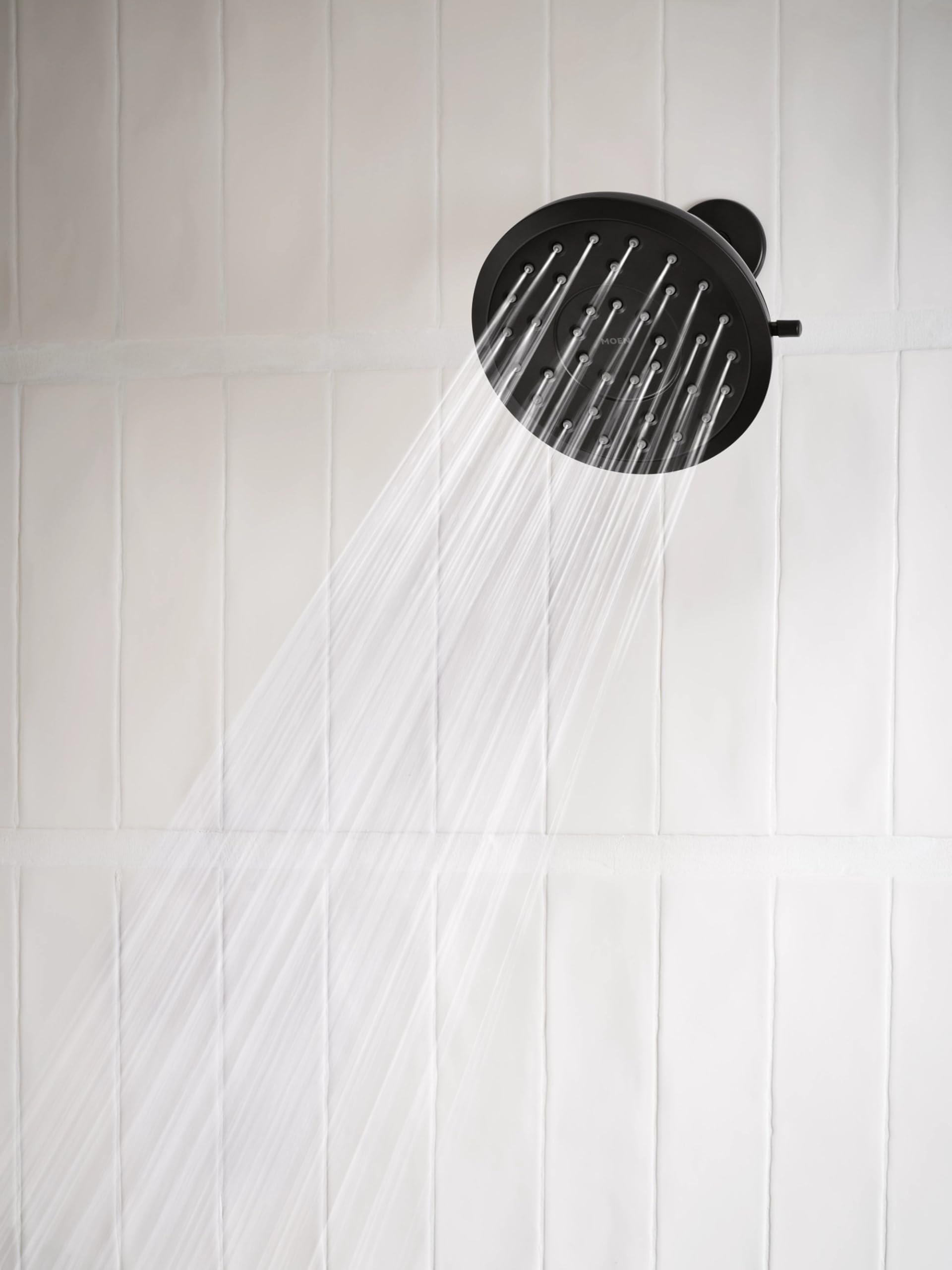 Matte black round rain shower head with control dial mounted on white tile wall with water streaming downward.