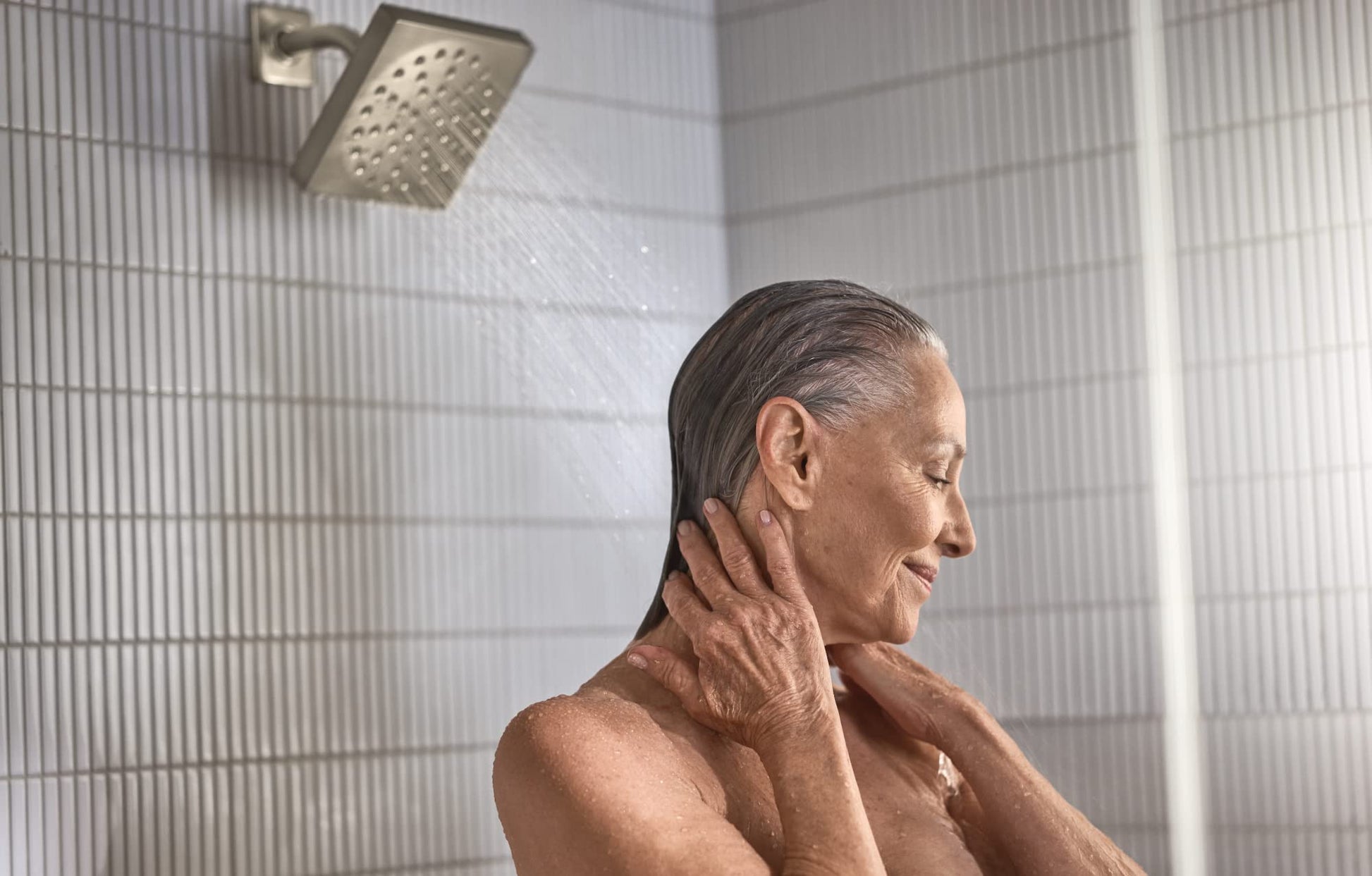 Brushed nickel square showerhead mounted on wall with water flowing over person's head in shower.