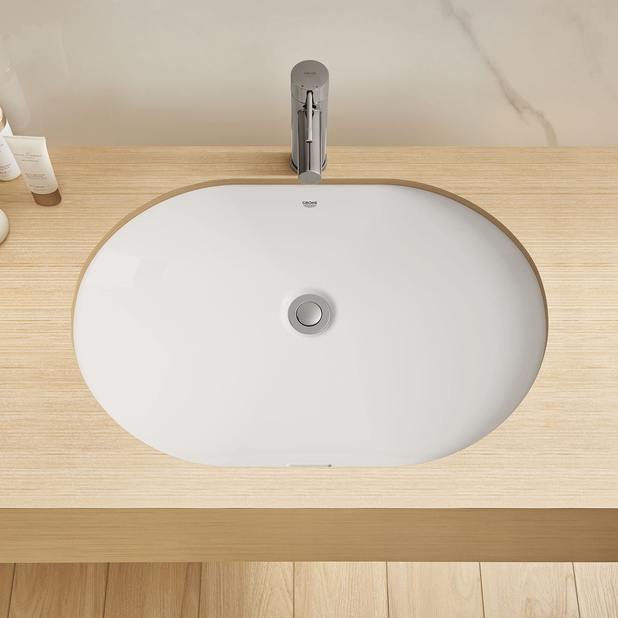 White oval undermount bathroom sink with chrome drain and faucet mounted on light wood countertop.