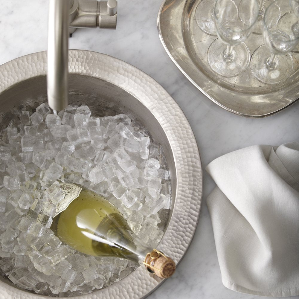 Brushed nickel bar sink with hammered white exterior filled with ice and a wine bottle, flanked by glasses and linens.