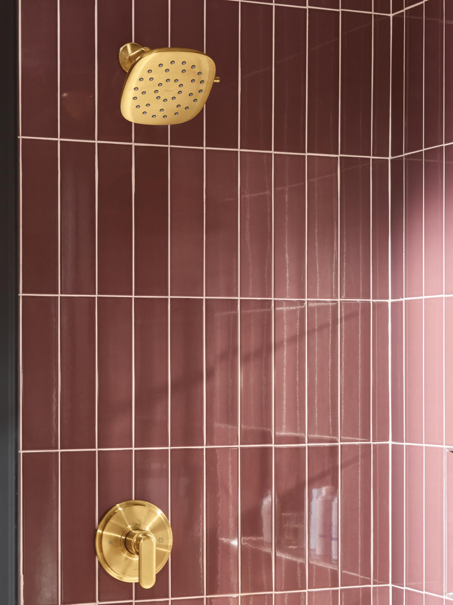 Square brushed gold rain shower head with multiple spray holes mounted on tile wall with matching round valve control below.