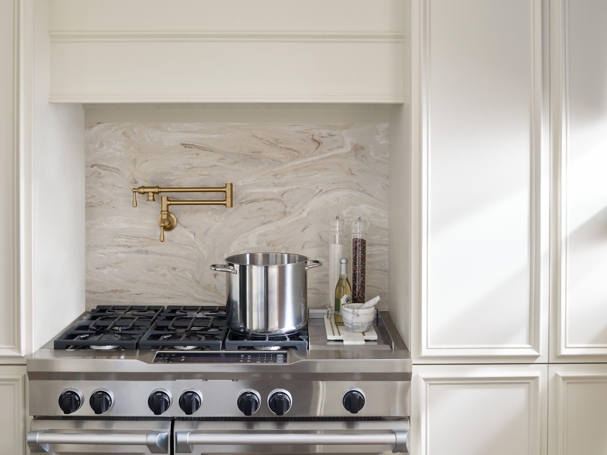 Brushed gold wall-mounted pot filler faucet with folding arm installed above a gas range with a stainless steel pot below.