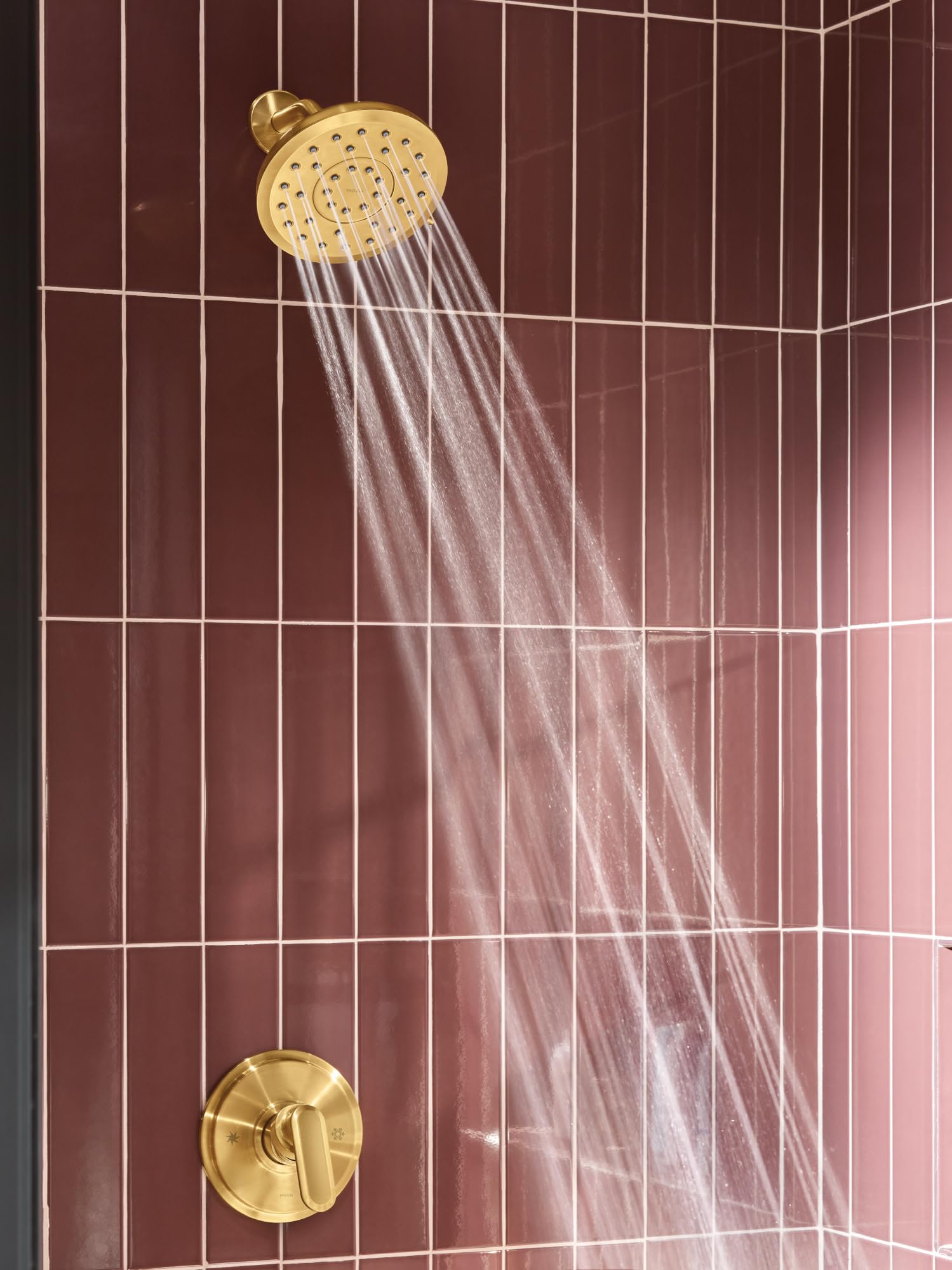 Brushed gold round shower head with spray streams, mounted on burgundy tiled shower wall with matching control dial below.