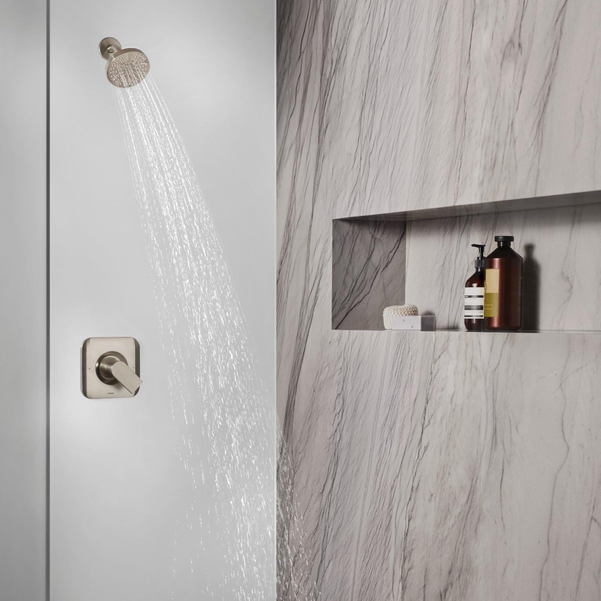 Brushed nickel shower valve trim with square control handle and round showerhead mounted on marble tile wall.