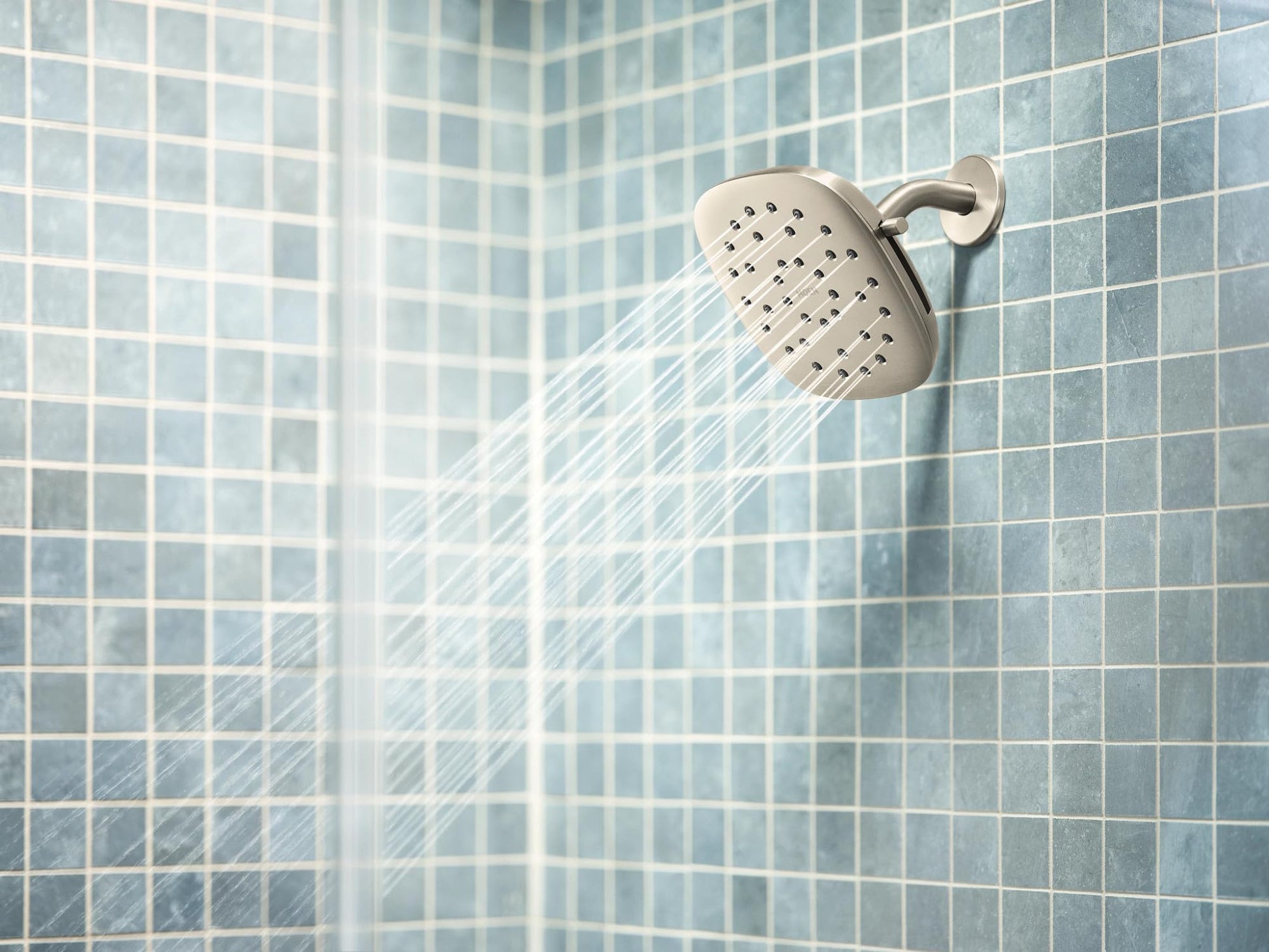 Square brushed nickel shower head with water spray, mounted on tiled shower wall.