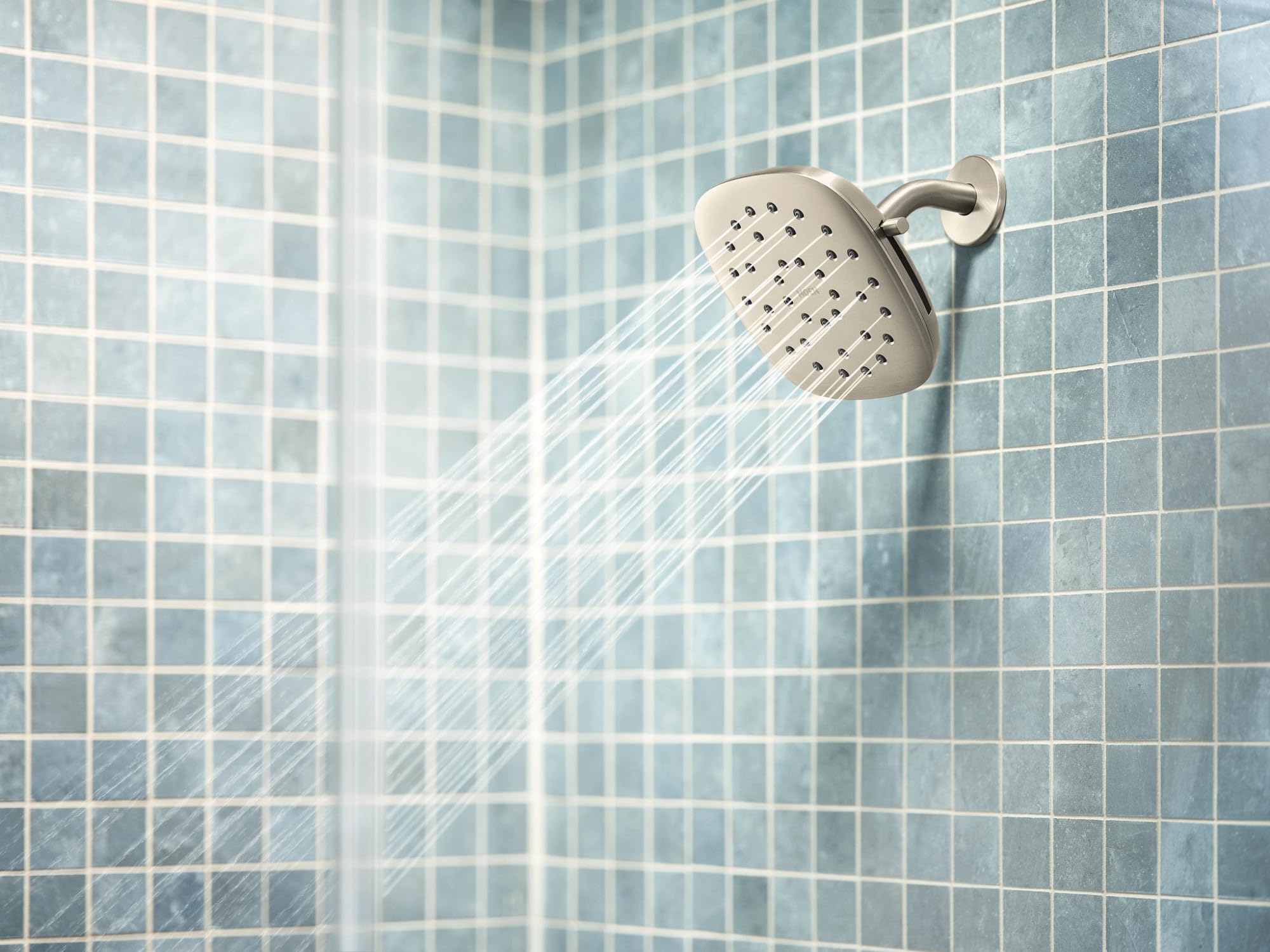 Square brushed nickel shower head with water spray, mounted on tiled shower wall.