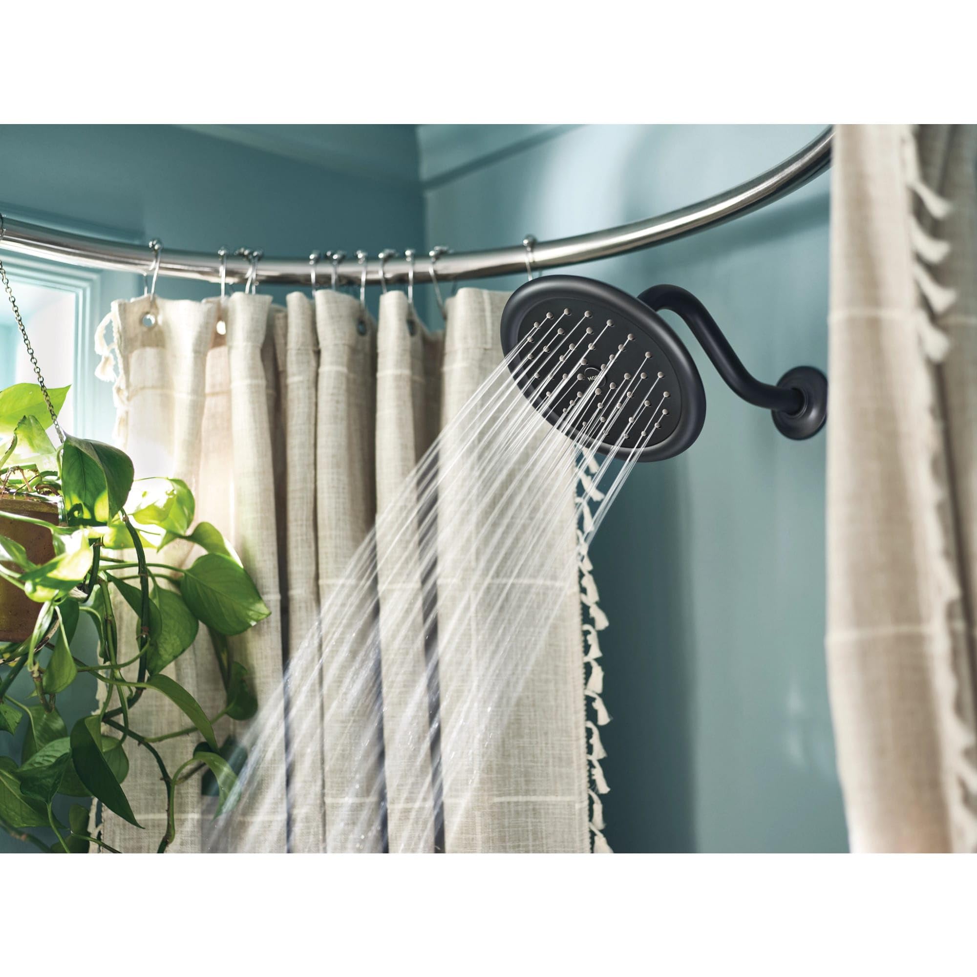 Black showerhead with water spray mounted on curved polished nickel rod above bathtub with beige curtain.