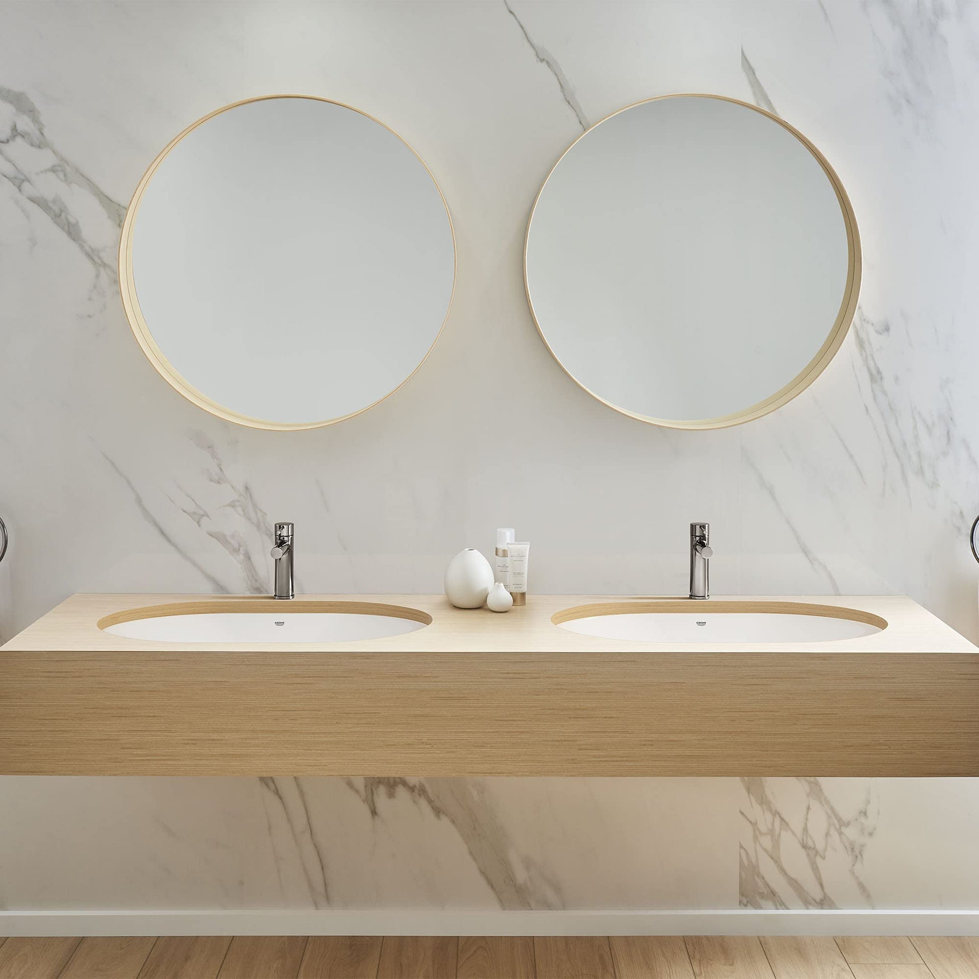 White undercounter bathroom sink with curved rim mounted in light wood vanity with two faucets.