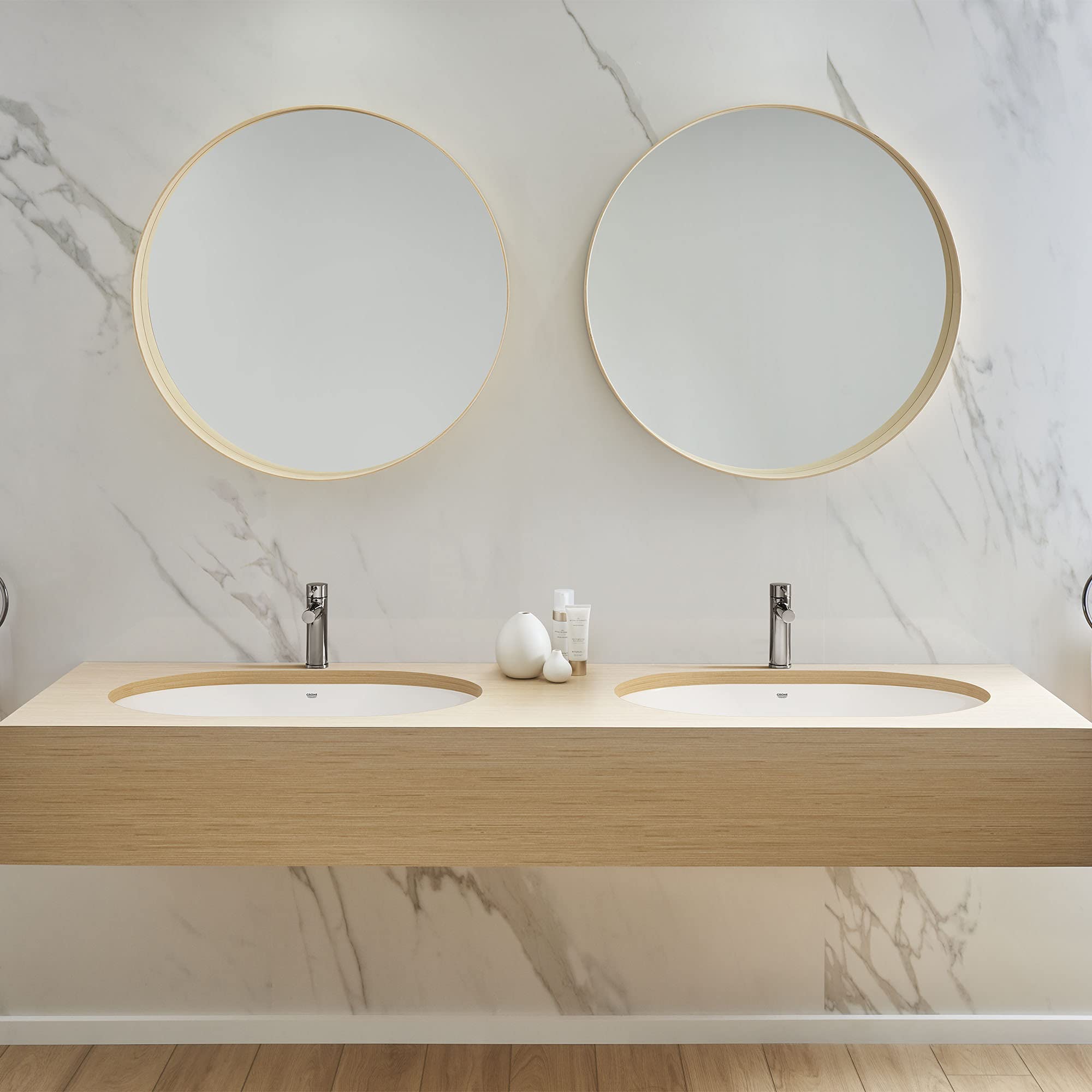 White undercounter bathroom sink with curved rim mounted in light wood vanity with two faucets.