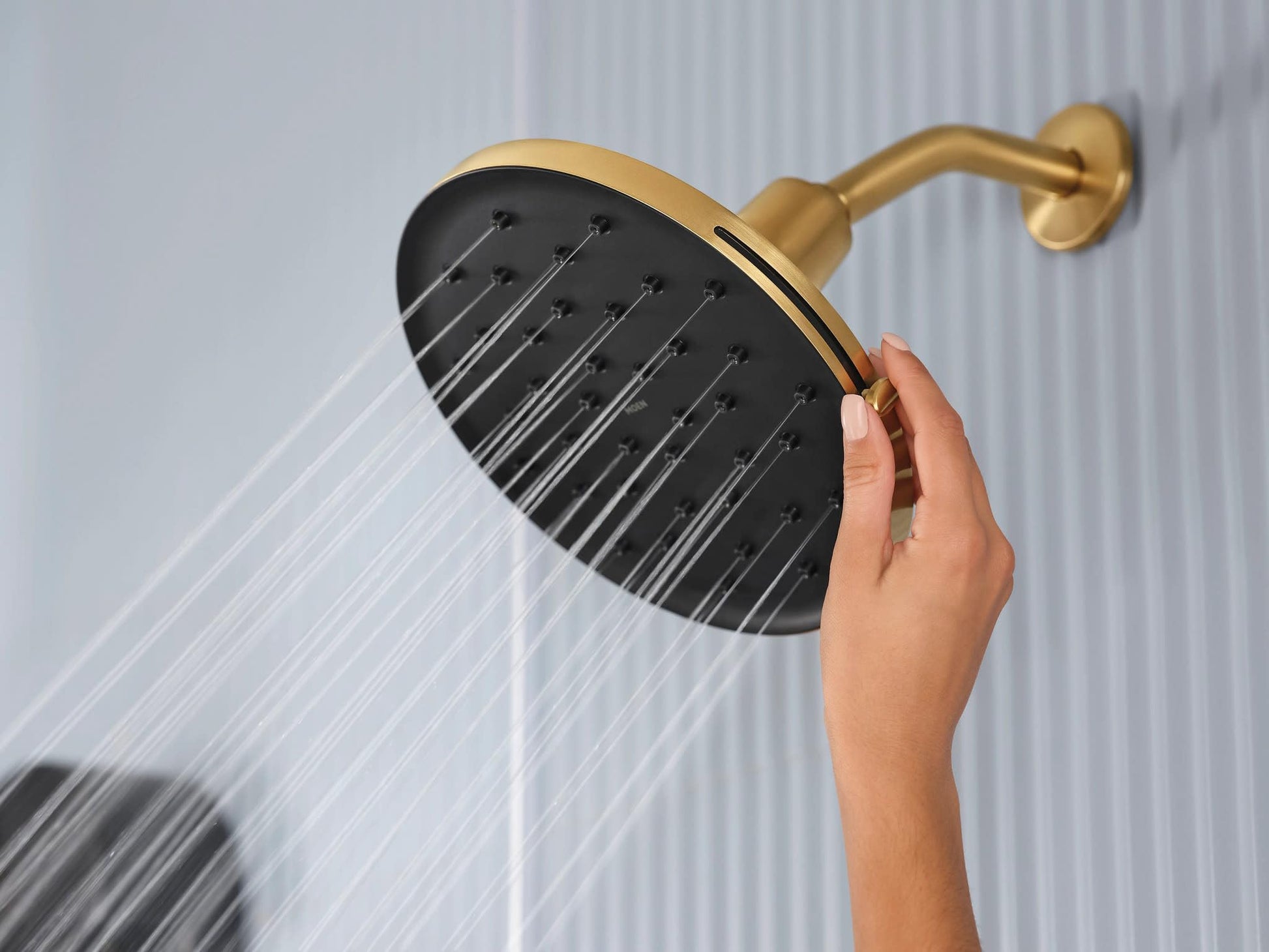 Brushed gold showerhead with black face and multiple spray holes, mounted on curved arm with water flowing downward.