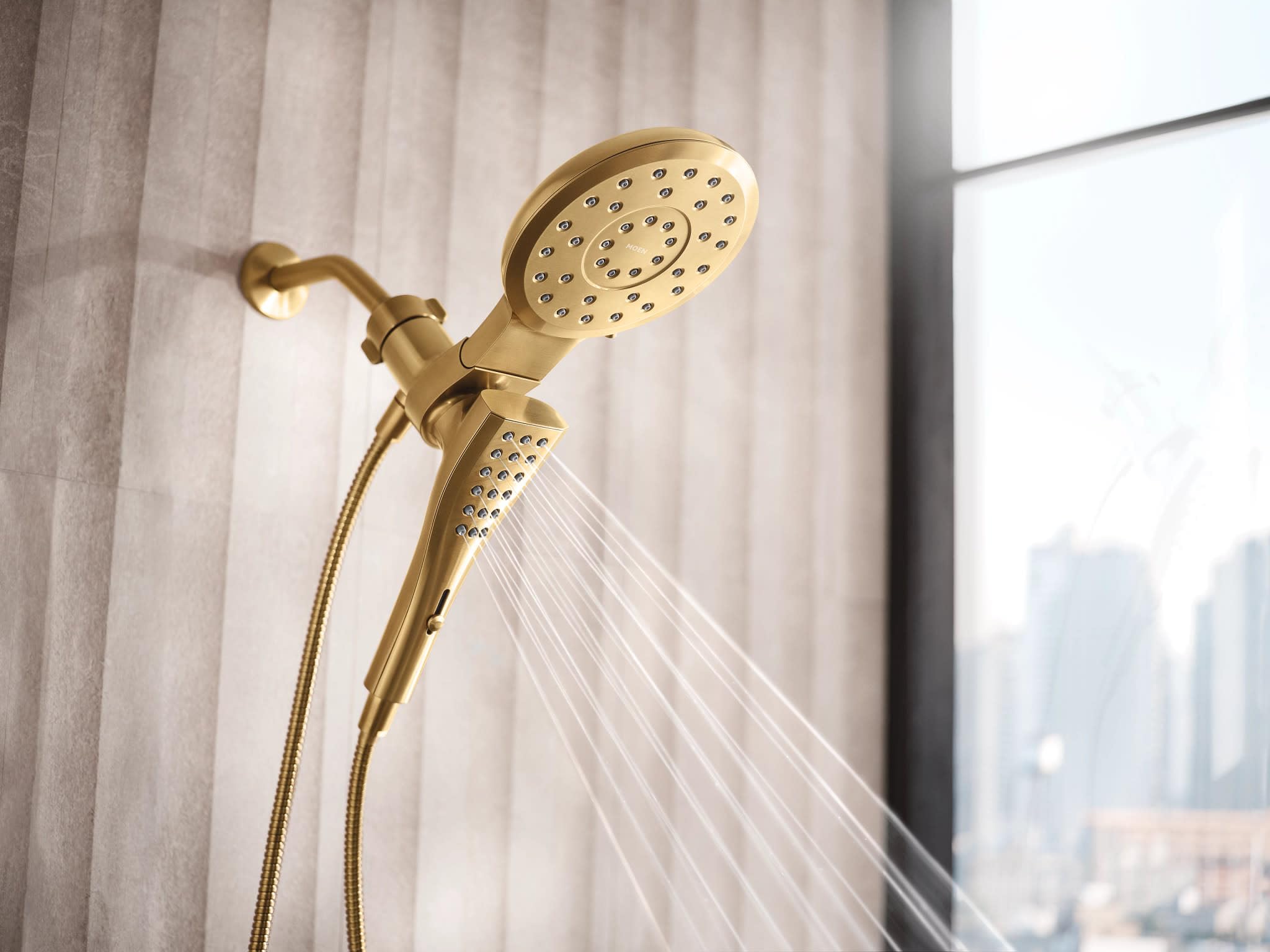 Brushed gold dual shower head system with upper round head and lower handheld wand mounted on wall with hose.
