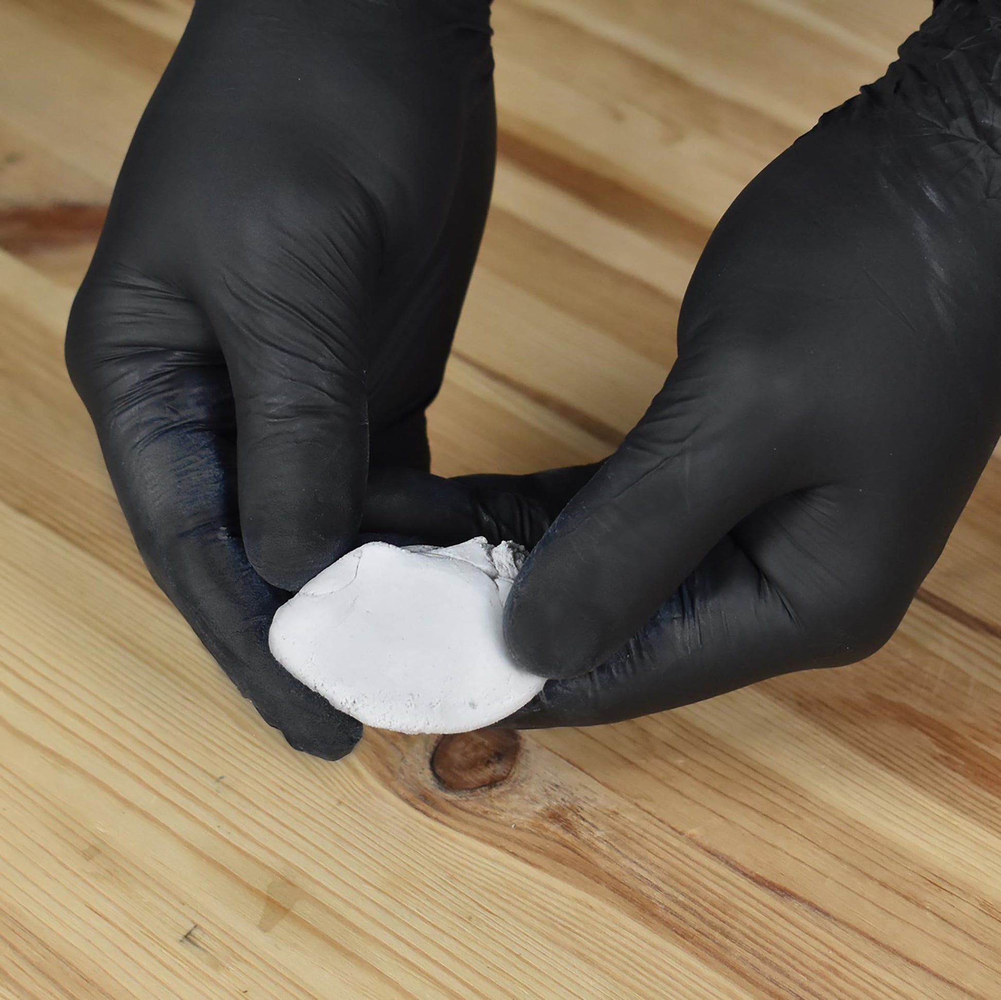 Gloved hands holding gray epoxy putty over a wooden surface with a hole.