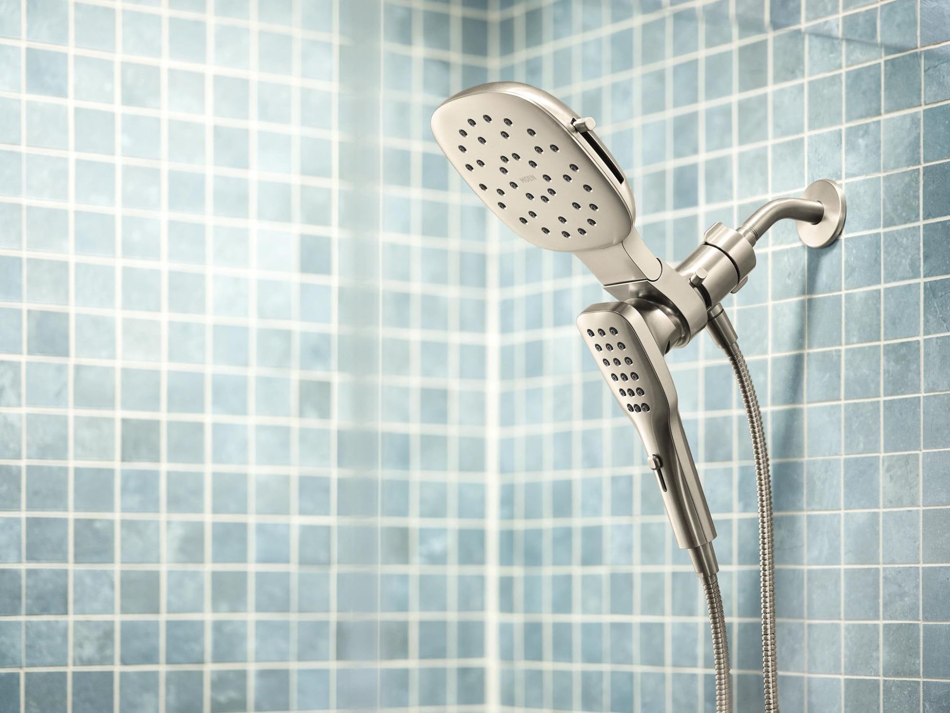 Brushed nickel shower combo with square rain head and detachable hand shower on flexible hose mounted against tile wall.