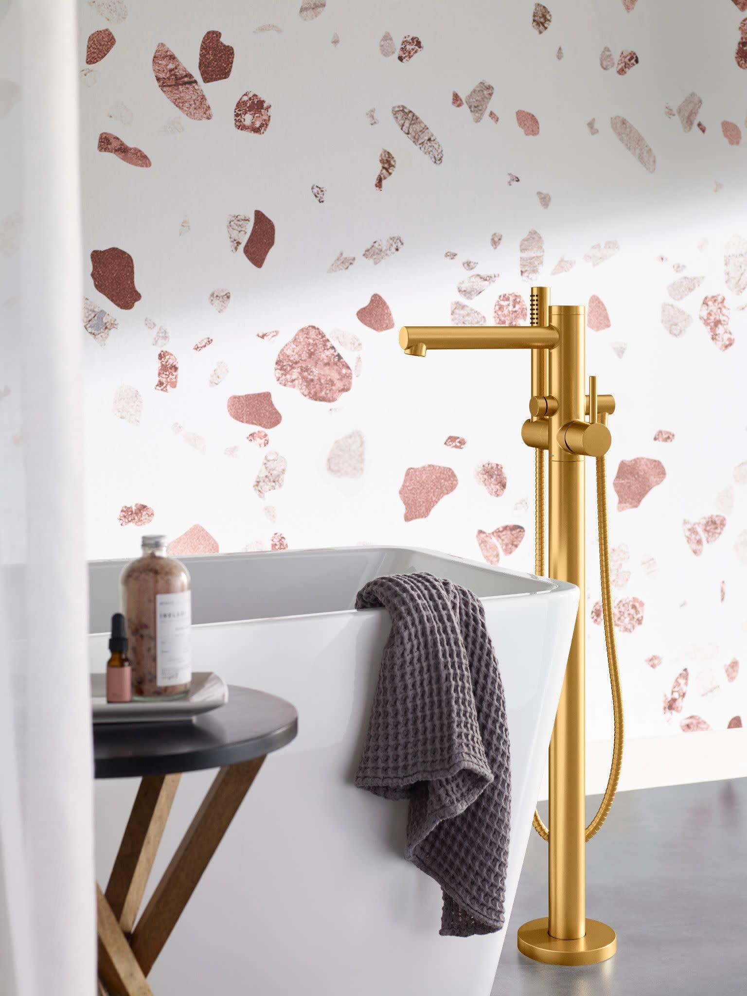 Brushed gold freestanding tub filler with single handle, spout, and attached handheld showerhead beside white soaking tub.
