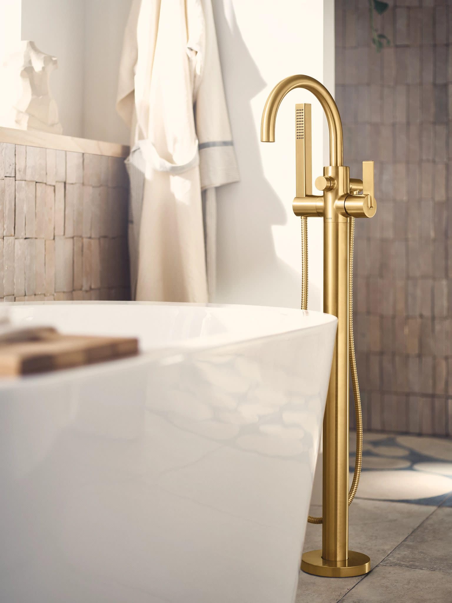Brushed gold freestanding tub filler with curved spout, single handle, and attached handshower hose next to white soaking tub